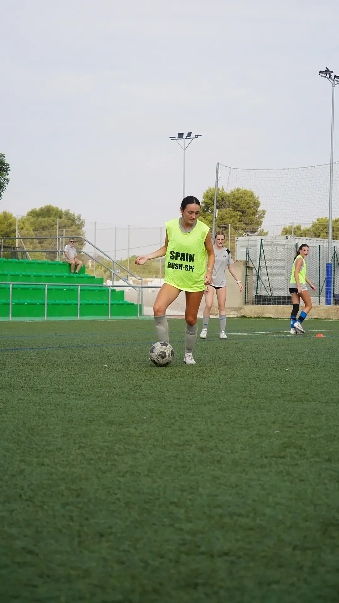 Soccer player in neon yellow vest dribbles a ball on a field, with another player in the background.