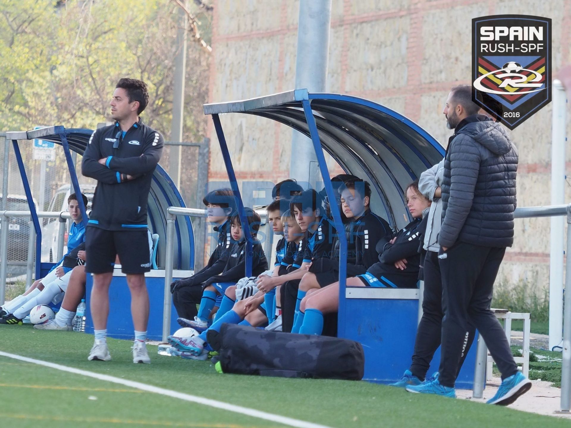Soccer team on a bench by the field, with coaches standing nearby during a match.