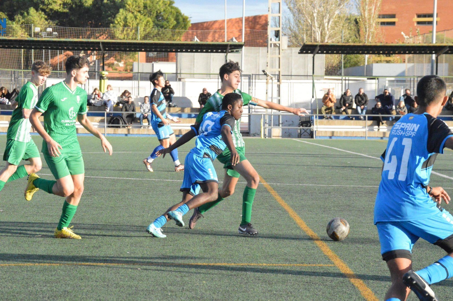 Soccer players in blue and green chase the ball on an outdoor field during a match