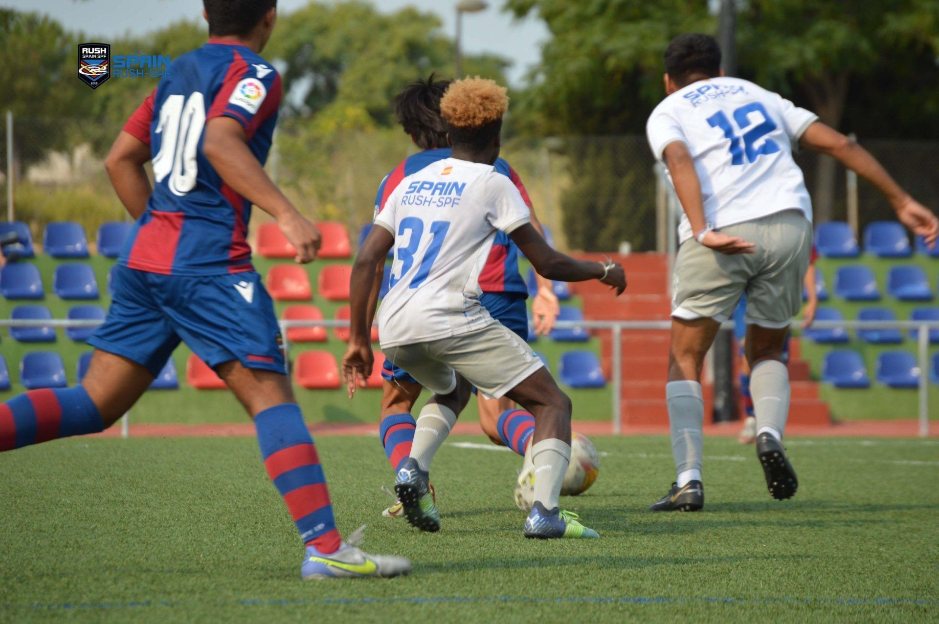 Soccer players in blue and white jerseys sprint toward the ball on a grass field