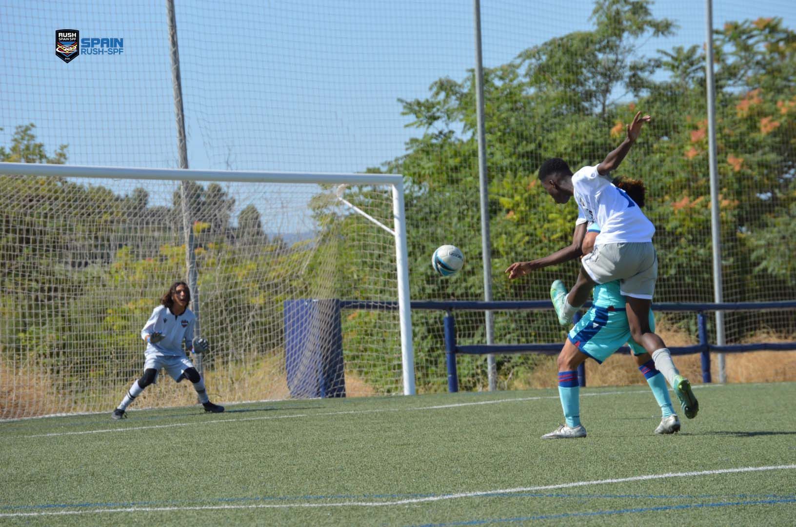 Soccer players in white and teal contest a ball near the goal on a sunny field