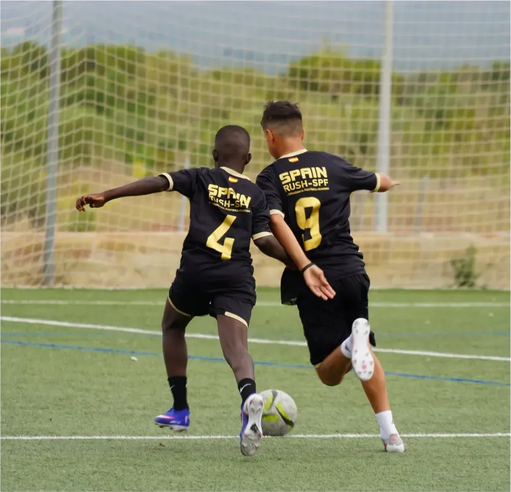 Two soccer players in black jerseys contest the ball on a field near the goal net.