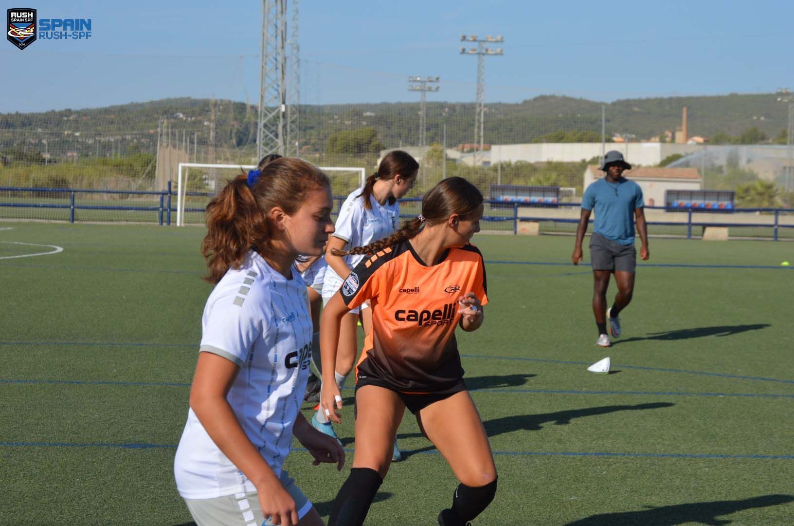 Youth soccer players in white and orange jerseys chase the ball on a sunny field.
