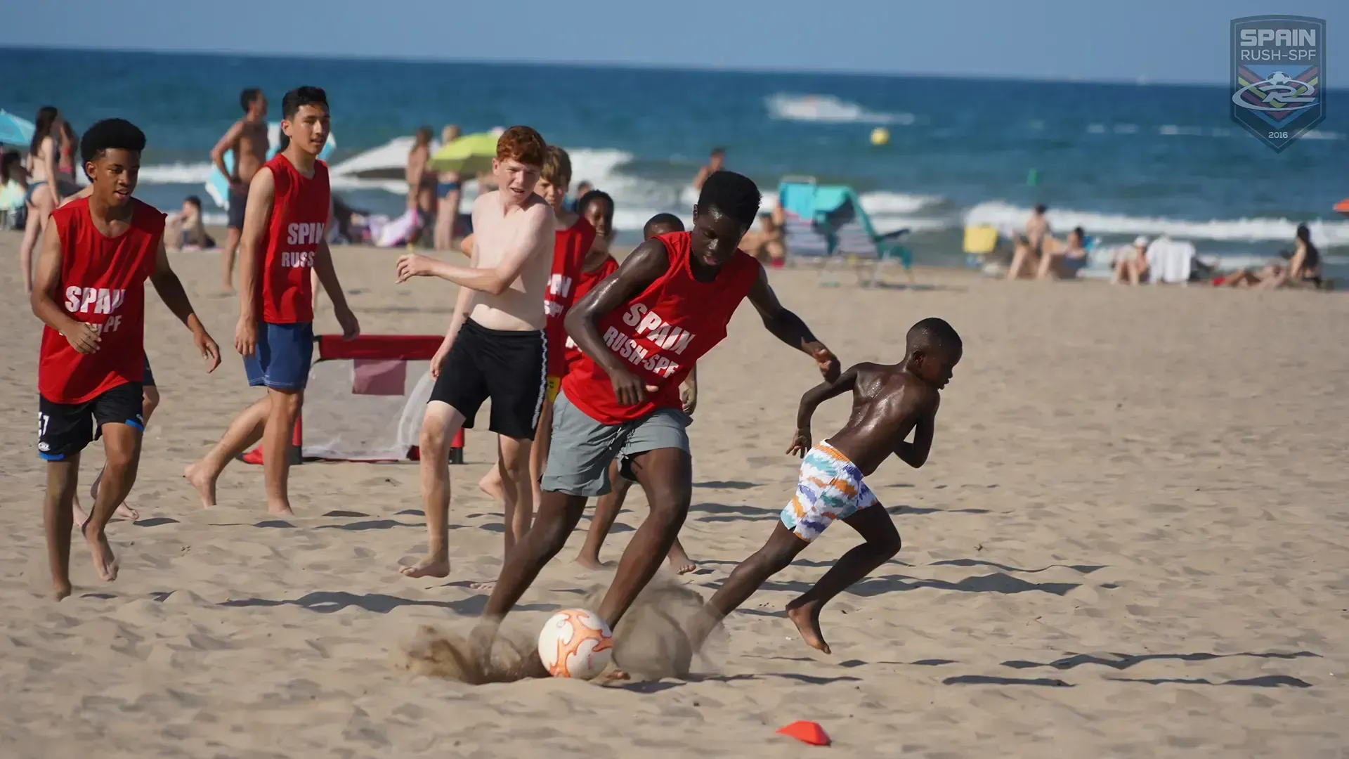 Soccer players in red jerseys chase a ball on a sunny beach near the ocean