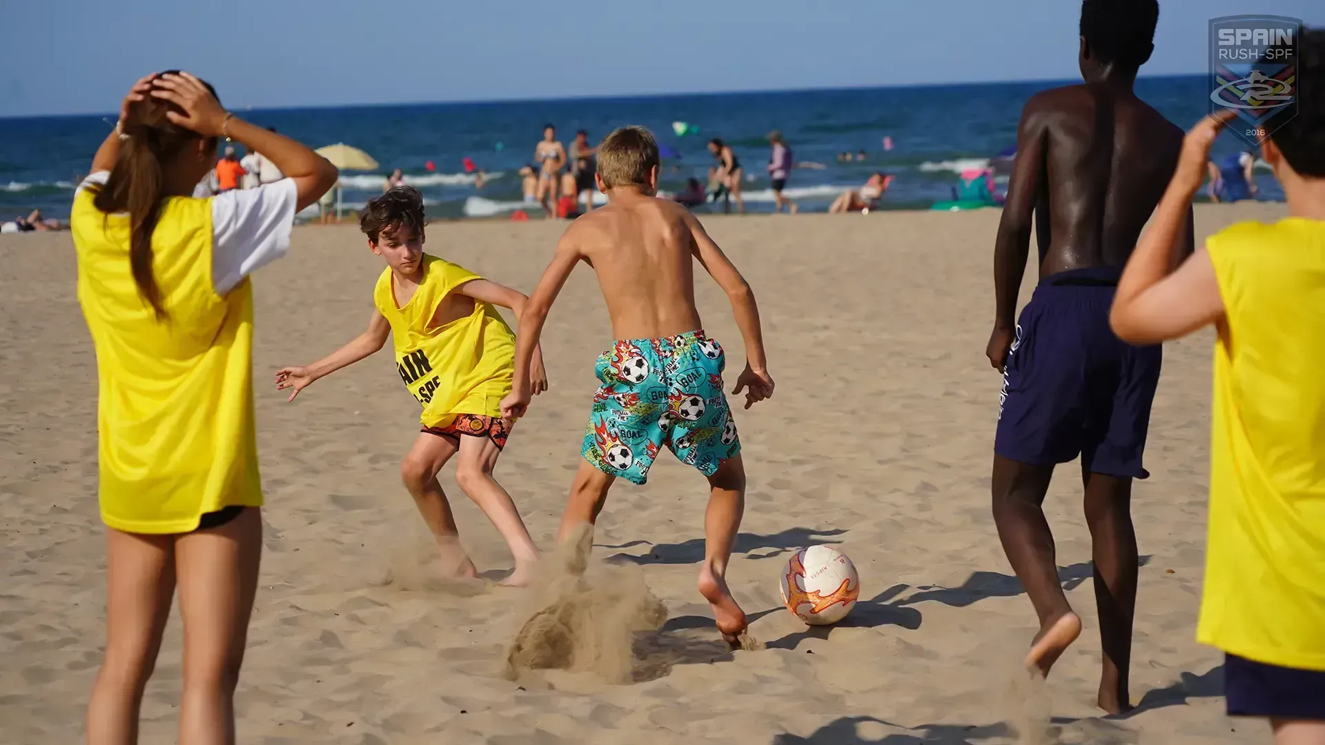 People playing soccer on a sandy beach near the ocean, with a ball mid-play.