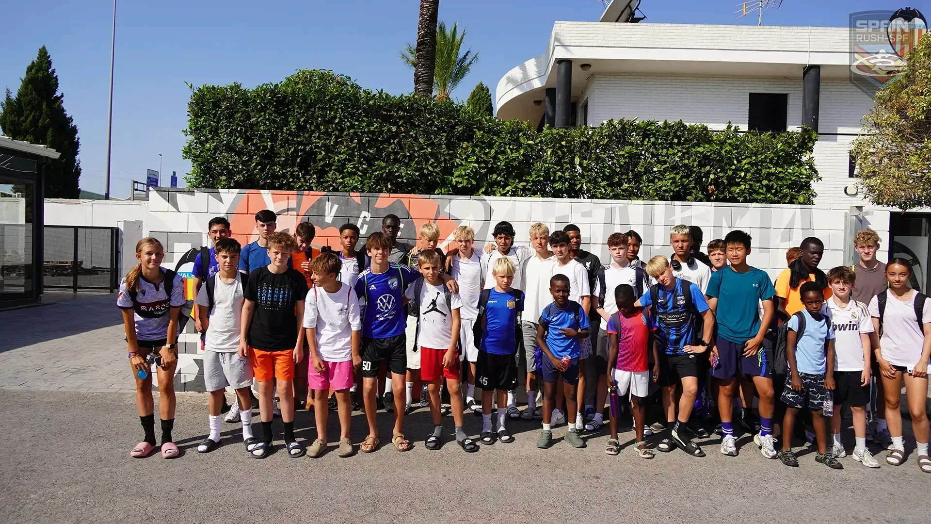 Group of children standing outdoors in sportswear, posing in front of a wall and house in bright sunlight