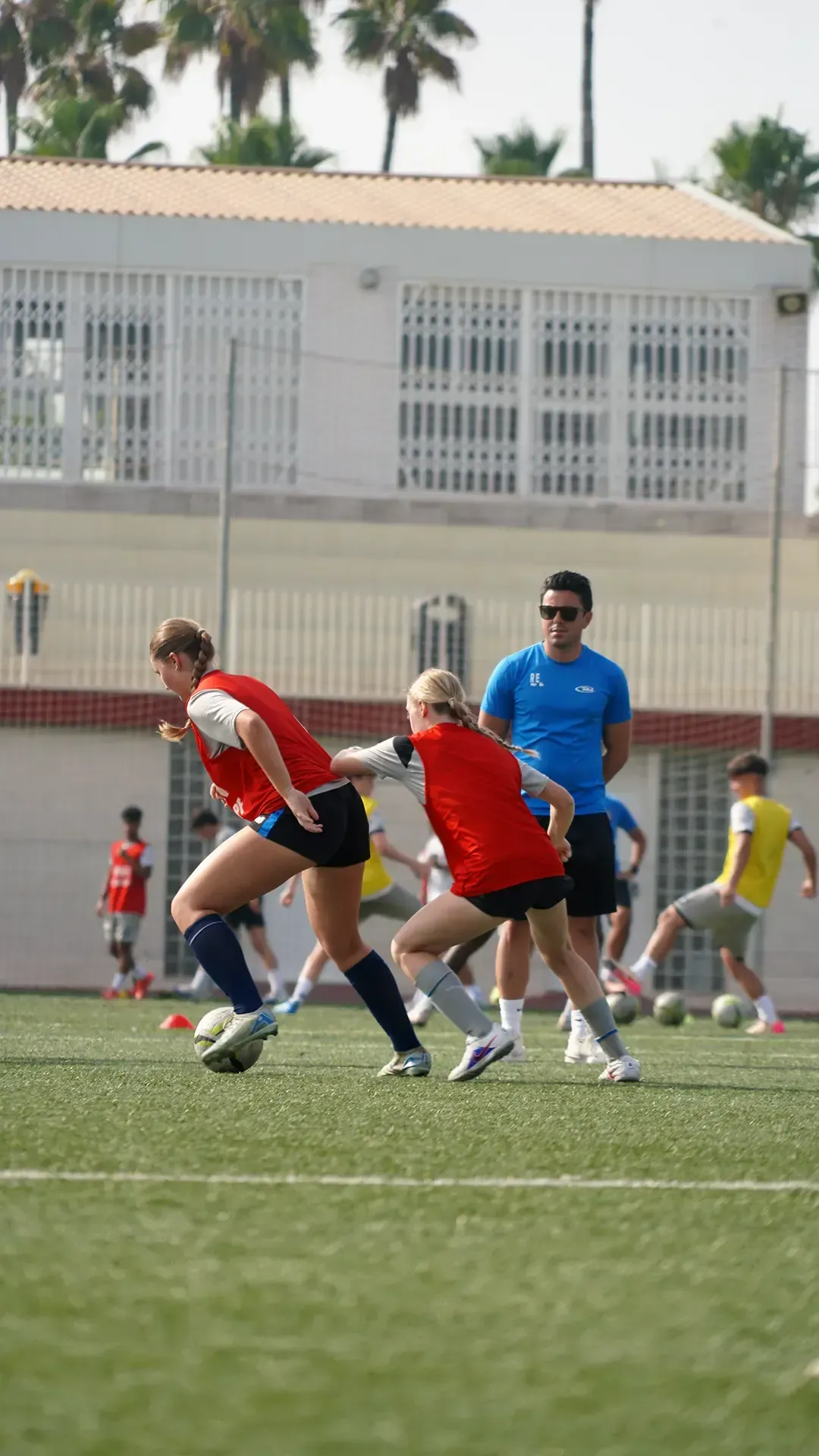 Soccer players in red and blue jerseys chase the ball on a field near a white building.