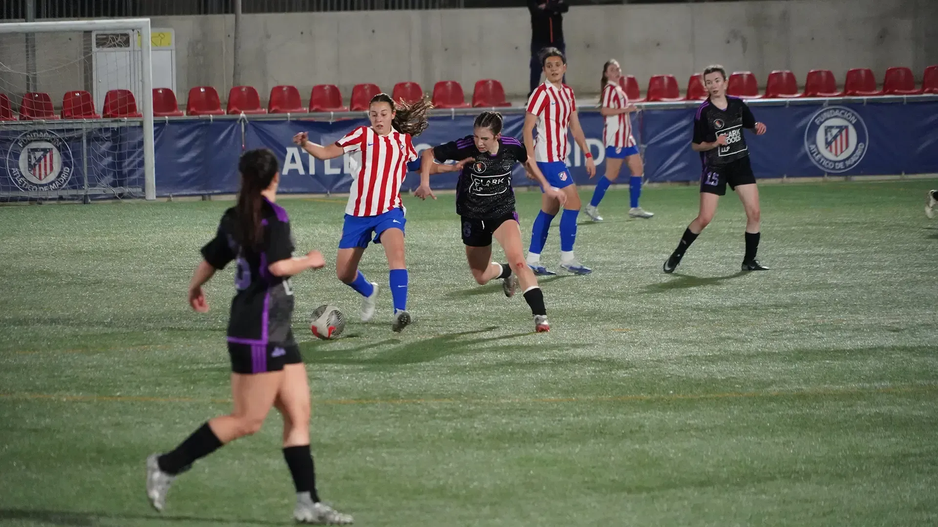 Soccer players in red-and-white and black uniforms compete on a wet field near empty red seats.