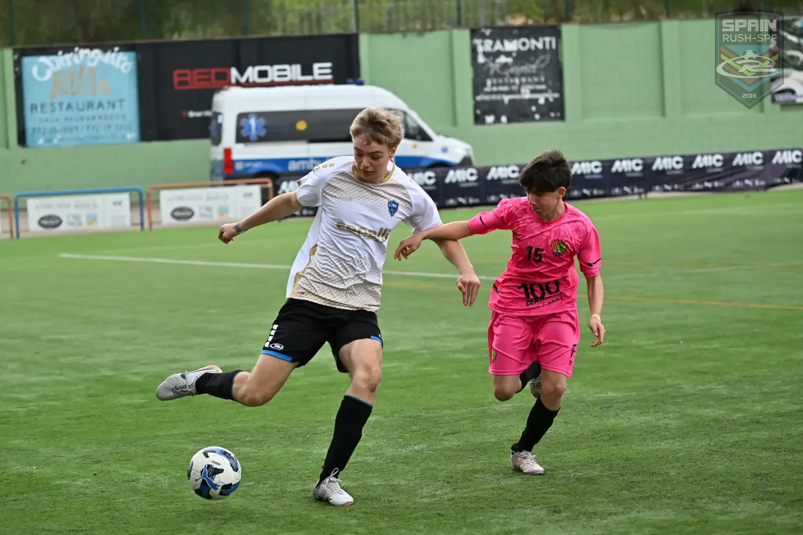 Two soccer players contest a ball on a wet field, one in white and black, one in bright pink.