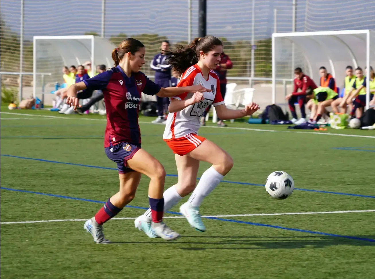 Two soccer players chase the ball on a grassy field near the goal during a match.