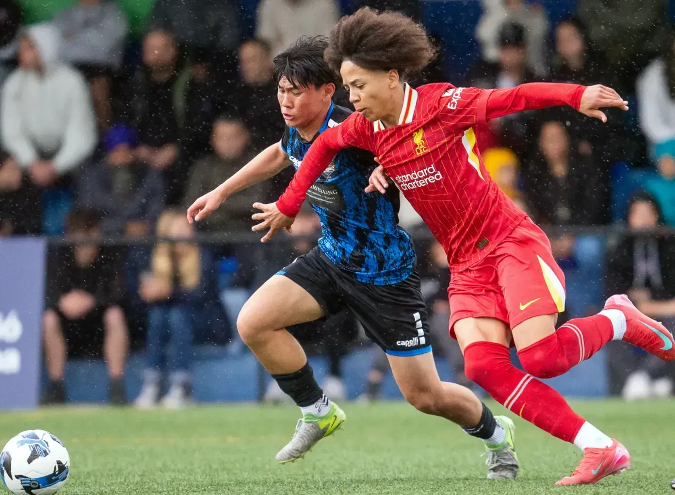 Two soccer players sprint after the ball, one in red and one in blue, during a match.