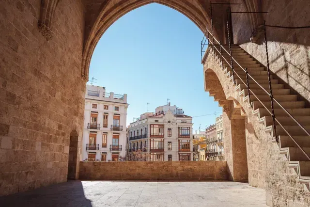 Stone archway framing a sunny plaza with historic buildings and an outdoor staircase on the right