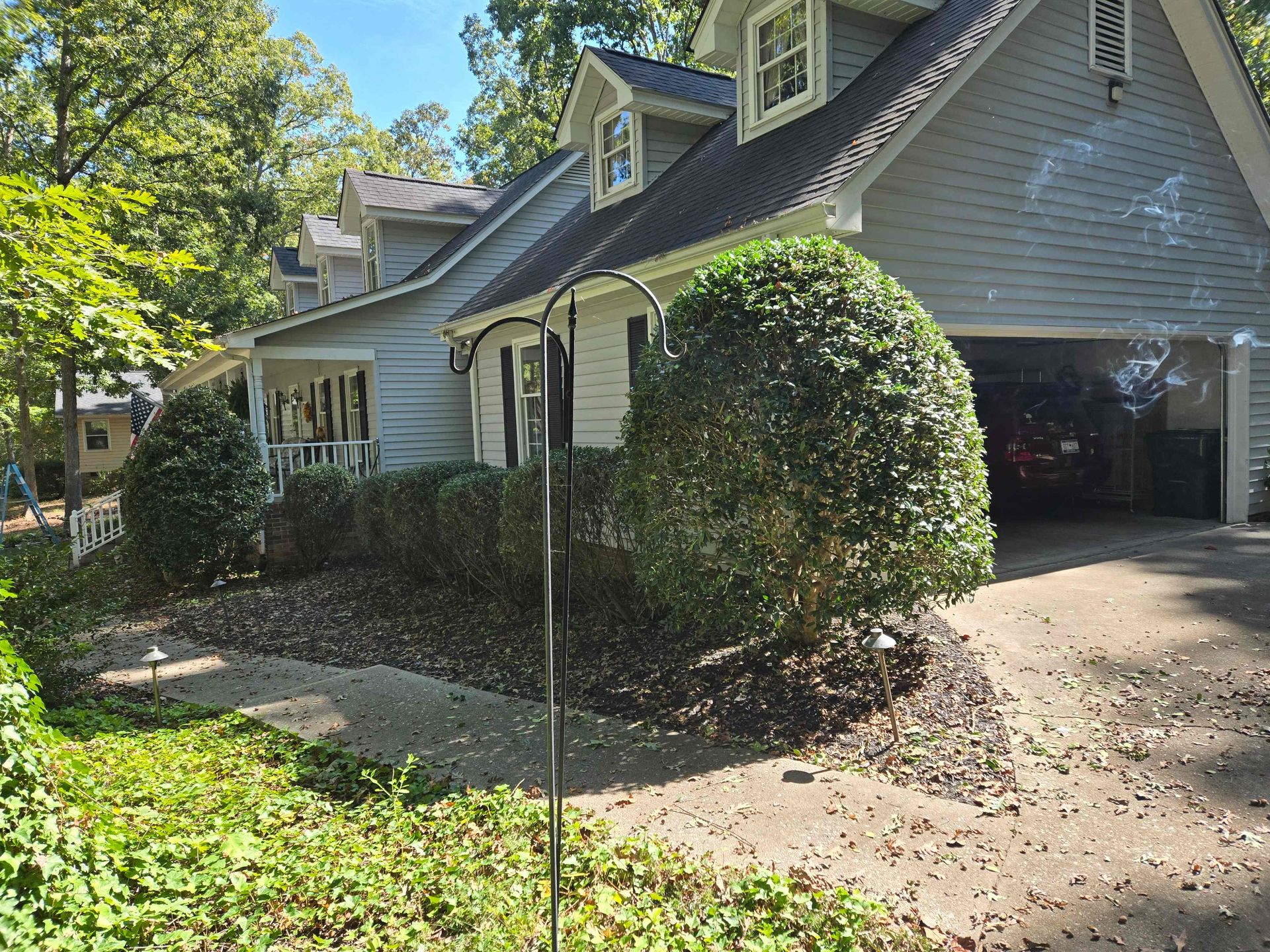 Suburban house with a driveway, garage, and large bushes beside a tree-lined yard