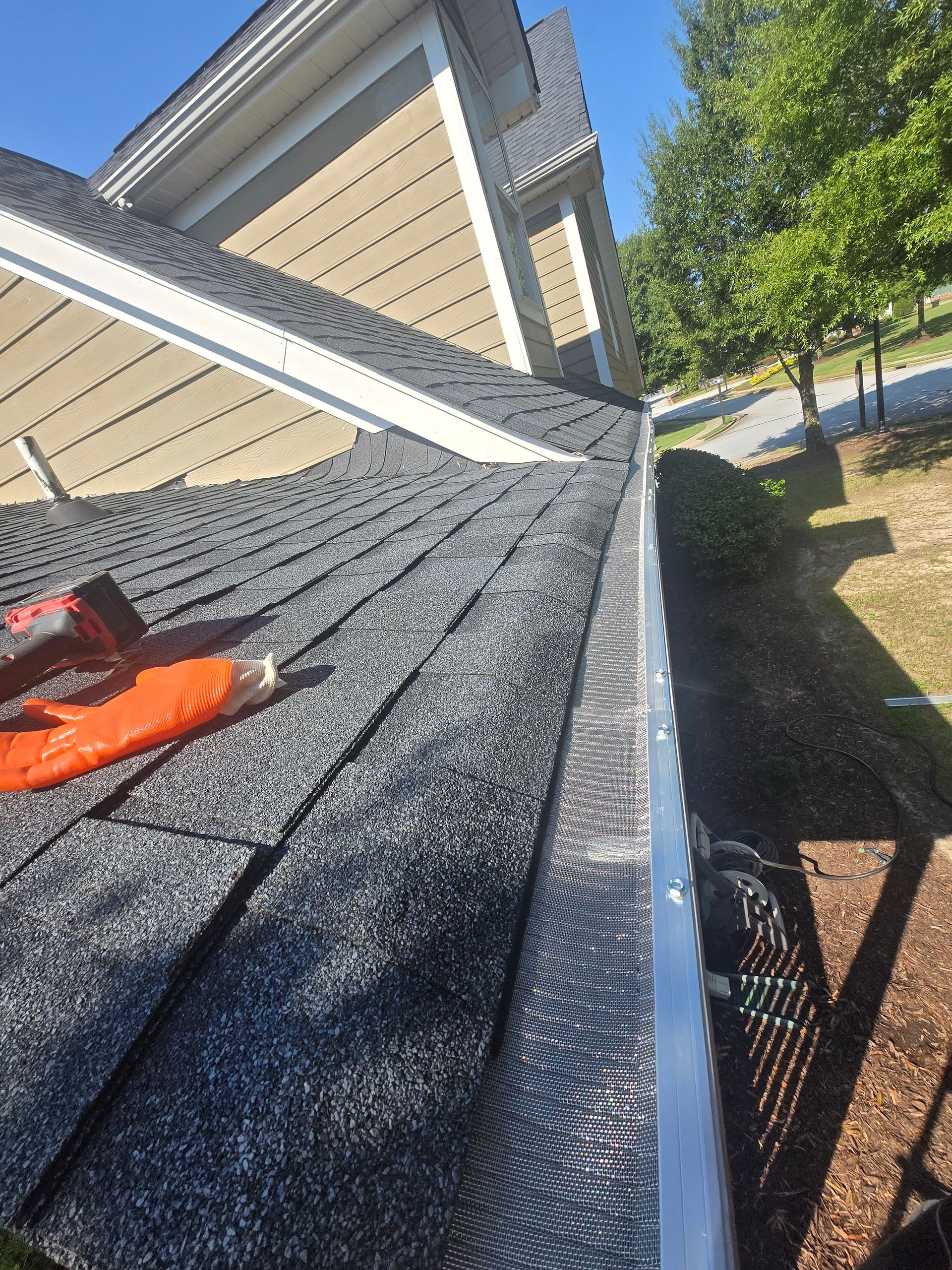 Roof shingles being installed beside a house, with an orange tool on the roof and a yard below.