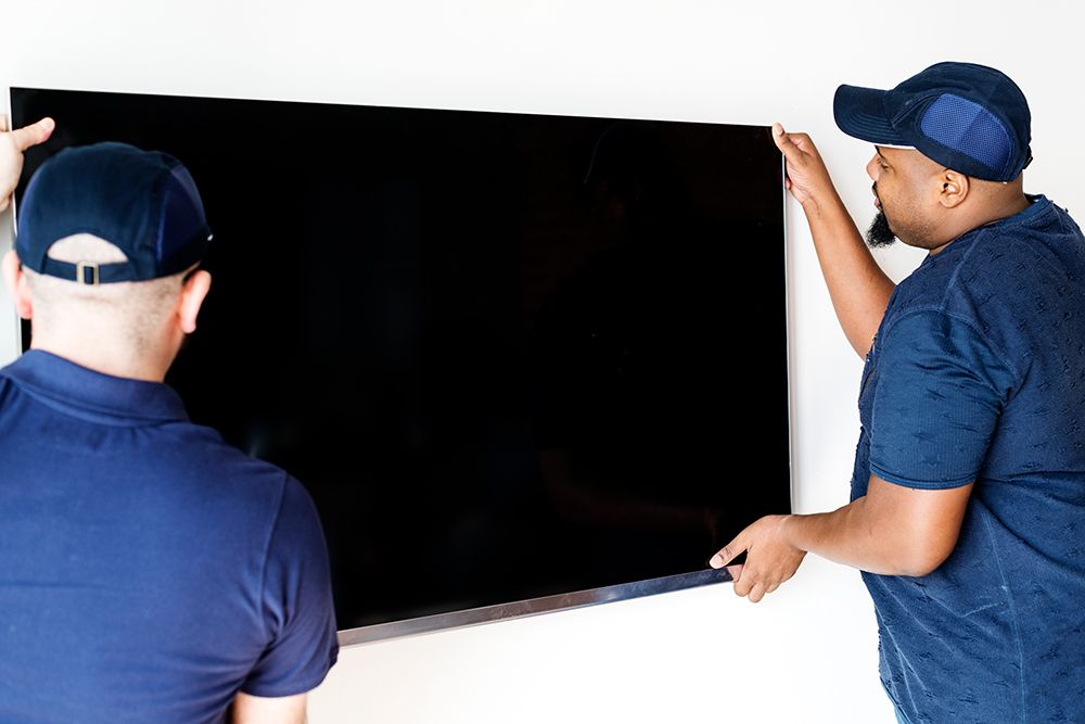 Two people in blue shirts and caps lift a large black TV, mounting it on a white wall.