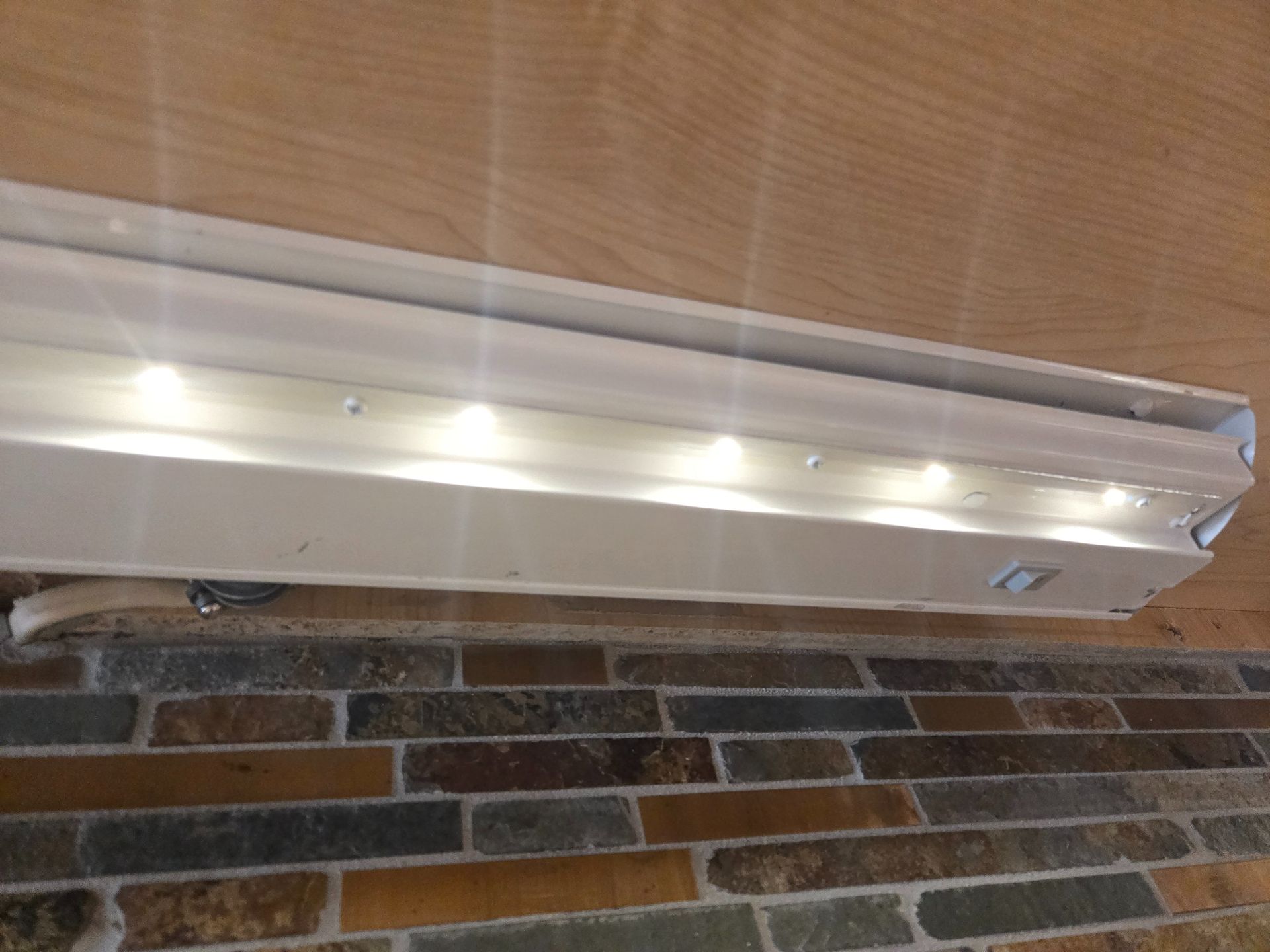 A white under-cabinet LED light strip mounted beneath a wooden surface, illuminating a multi-colored stone tile wall.