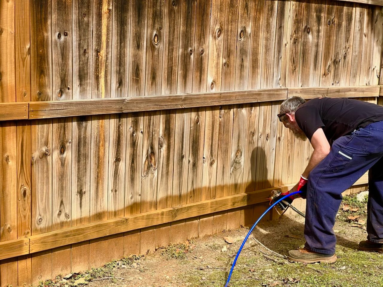 A person in dark clothing uses a sprayer to apply stain to a wooden fence outdoors.