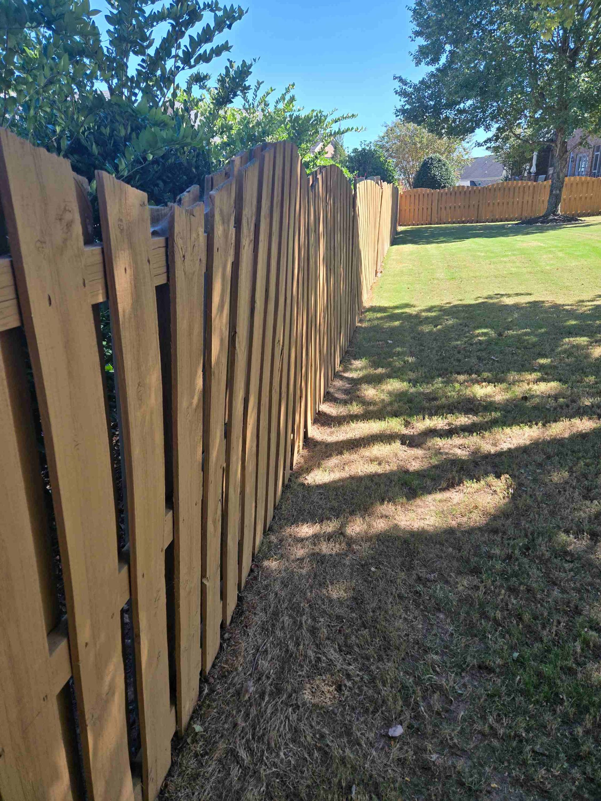 A long, weathered wooden picket fence lines a grass yard under a clear blue sky.