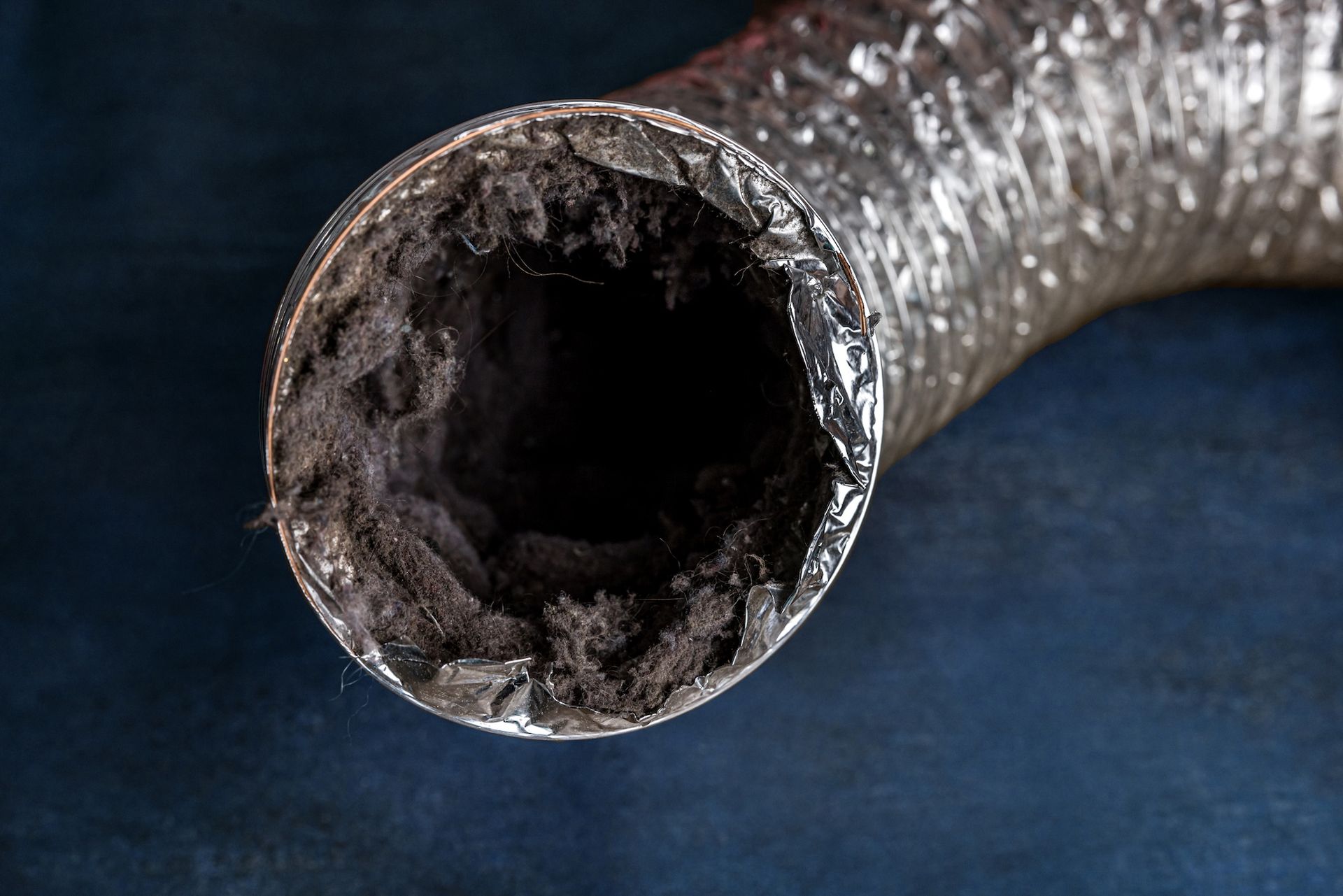 A close-up of a dusty, clogged silver flexible duct with thick, black debris inside.
