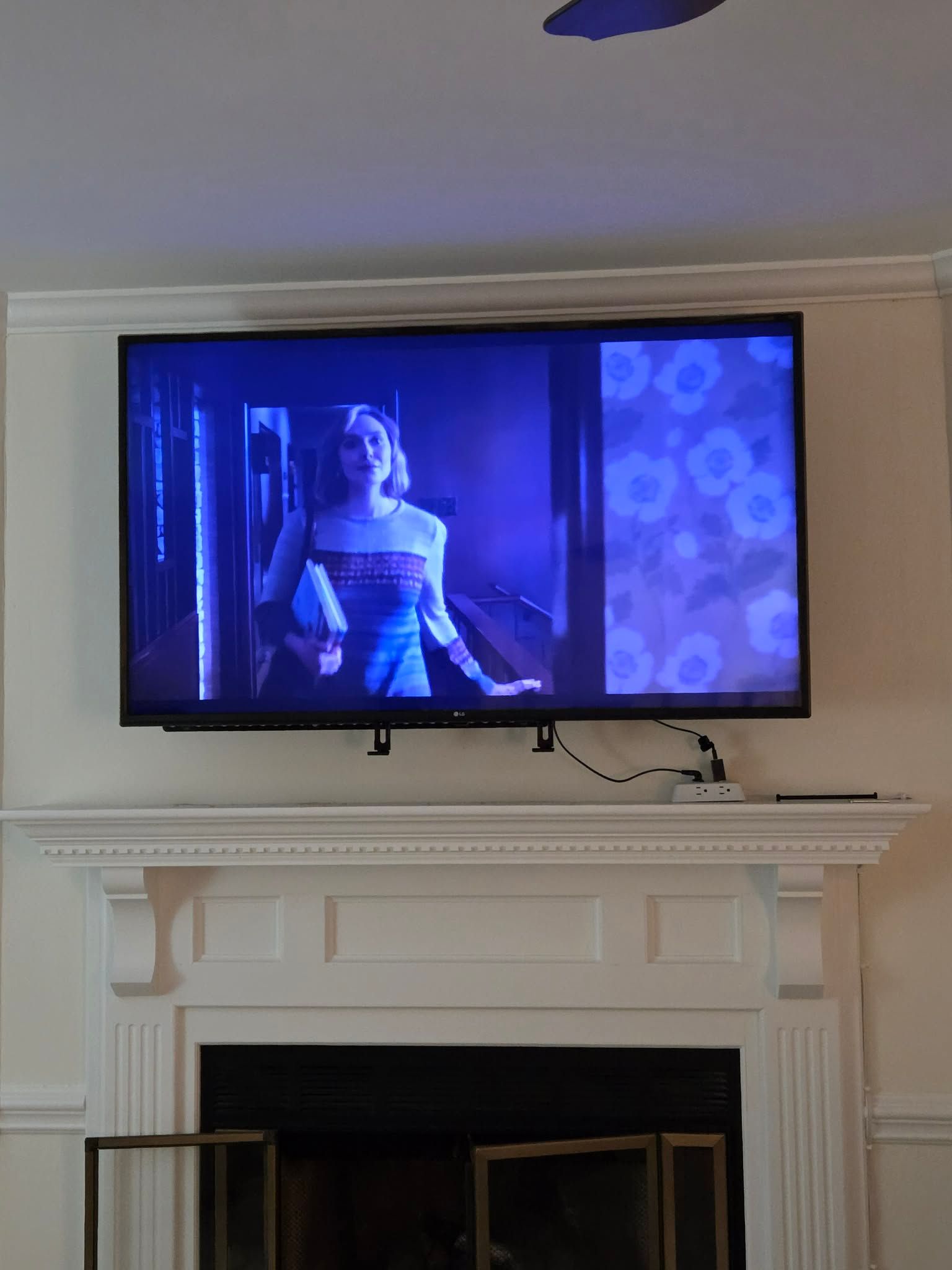 A wall-mounted TV above a white fireplace mantel displays a person on screen with a blue, flower-patterned side panel.