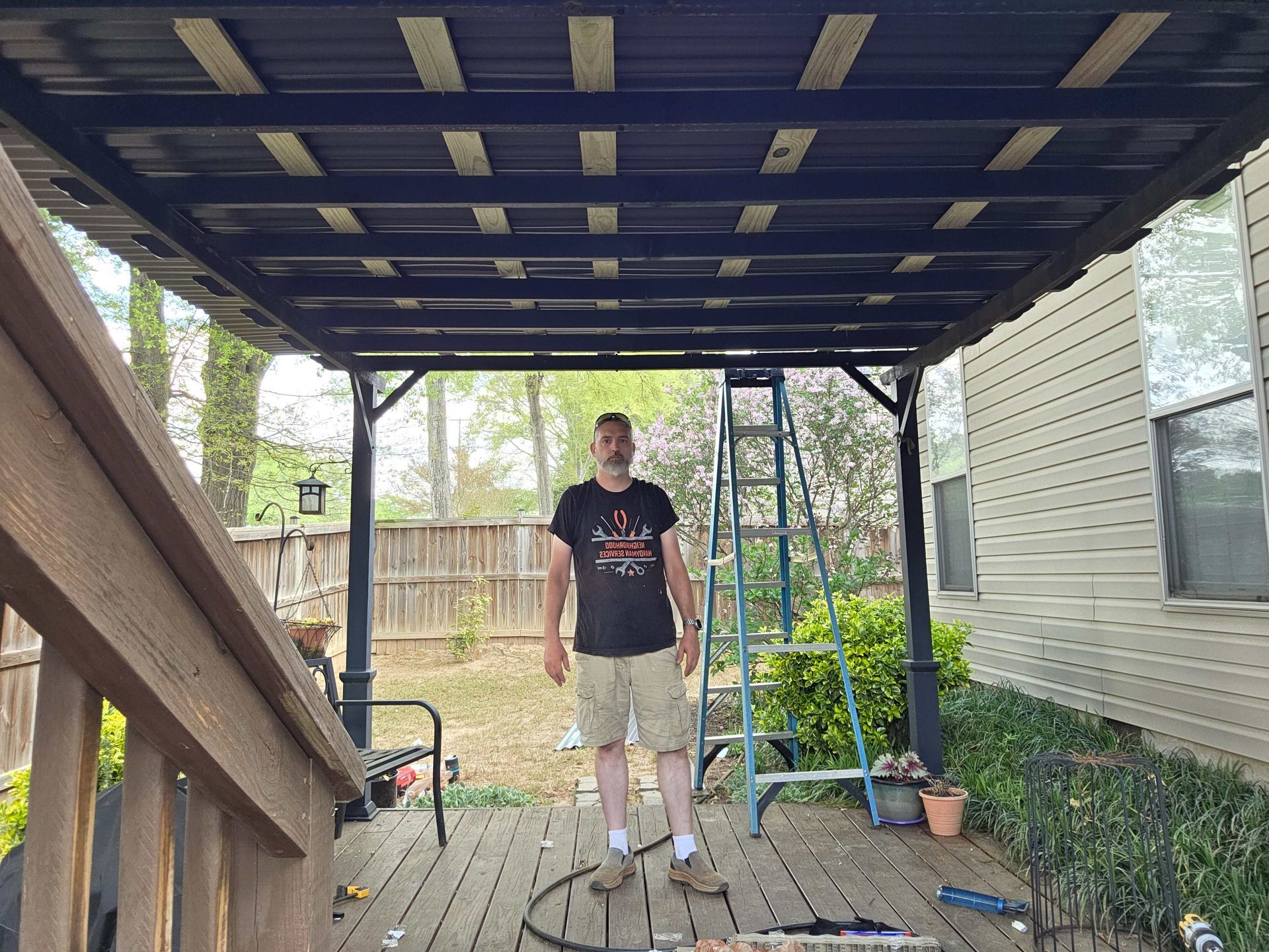 Person standing on a wooden deck under a blue canopy, with a ladder and house exterior nearby.