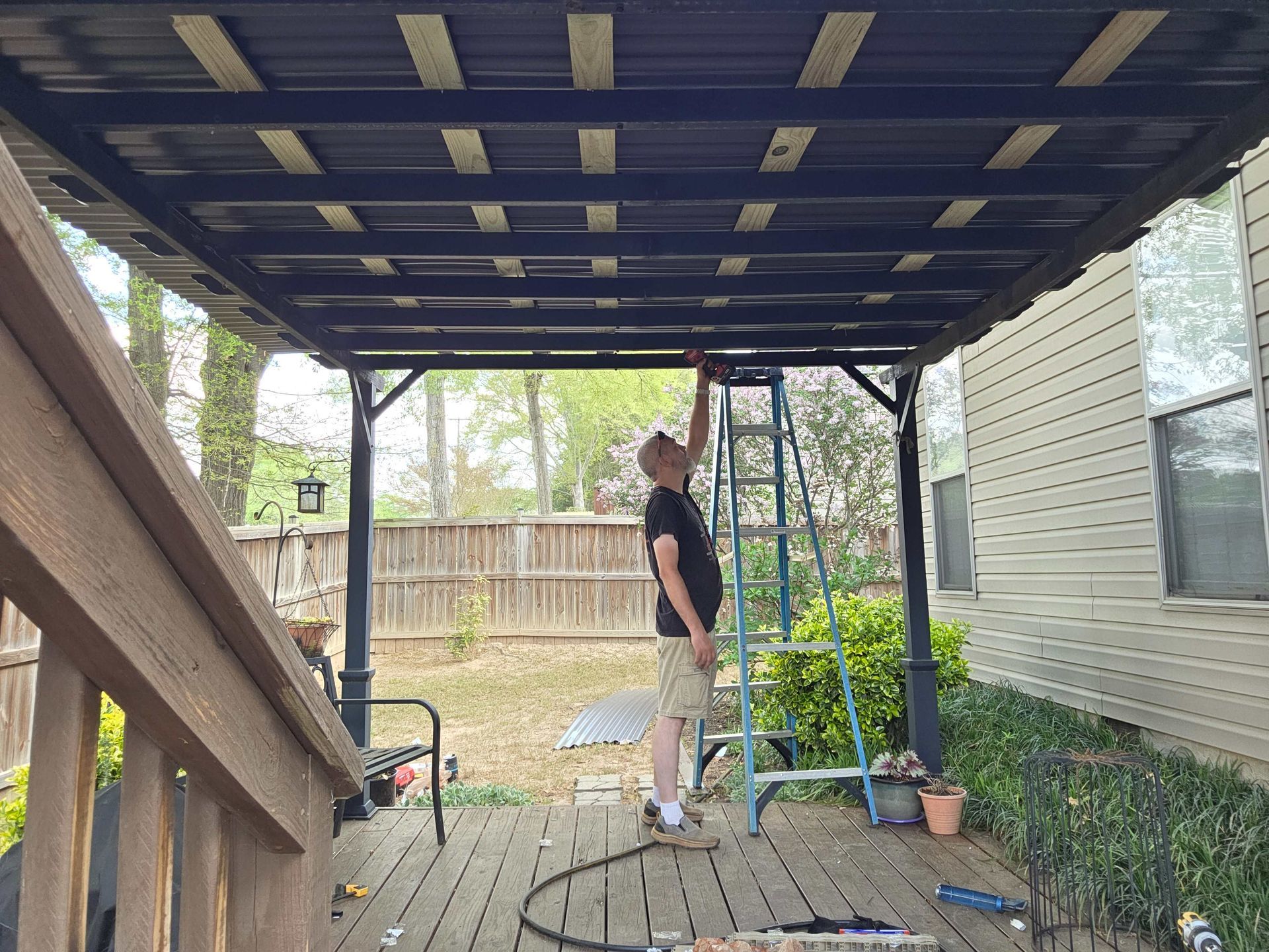 Person standing on a deck under a covered patio, working near a ladder beside a house and fence.