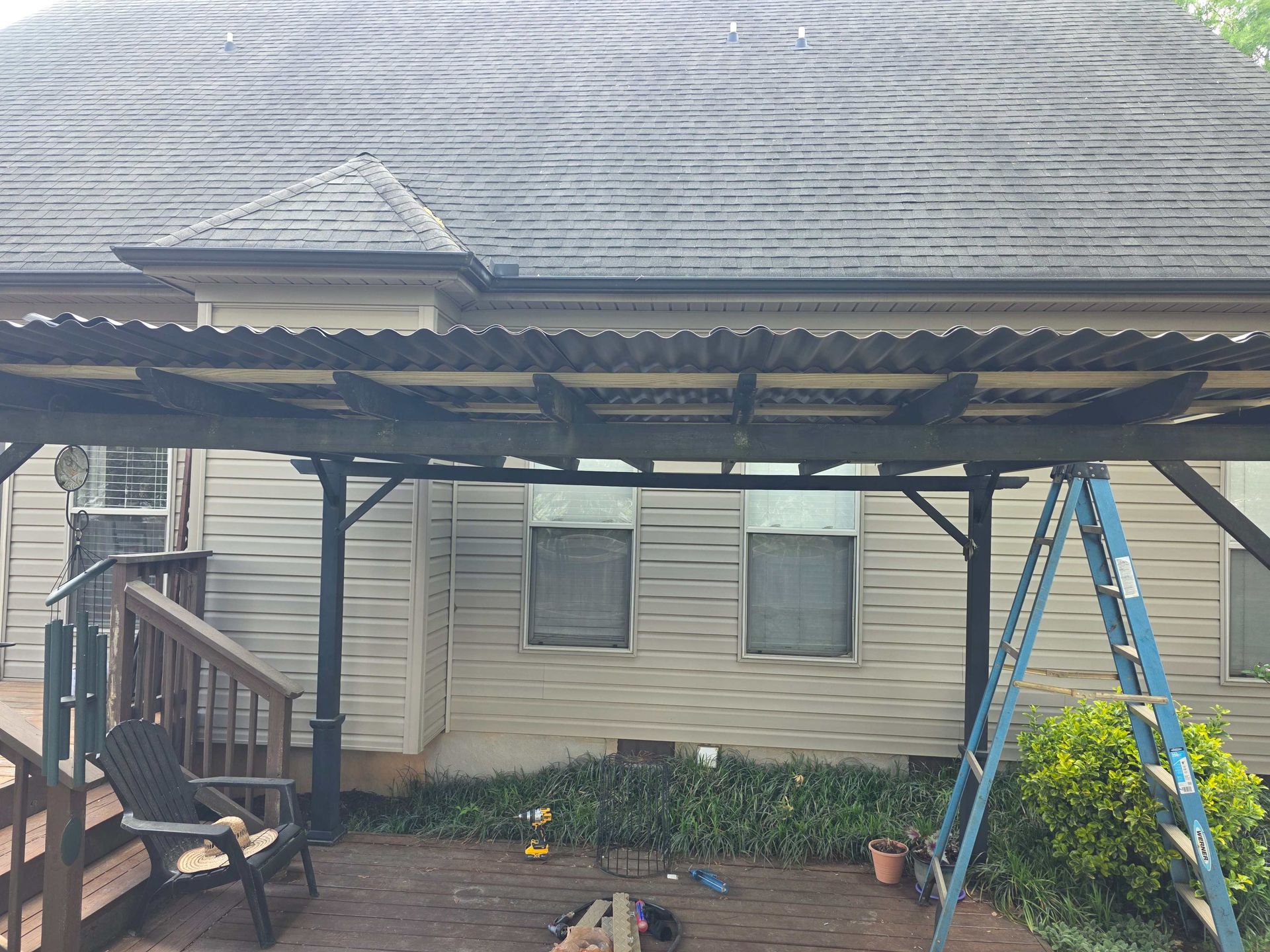 Covered backyard patio beside a beige house, with stairs, blue ladder, and a brick path.