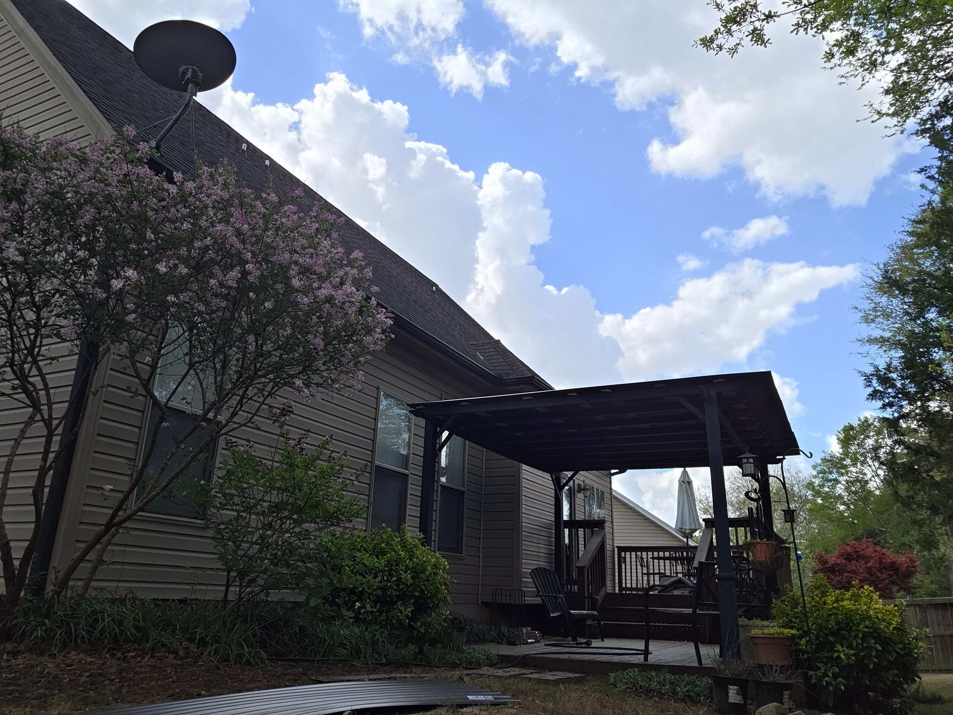 Patio beside a brick house with a covered seating area, shrubs, and a blue sky with clouds