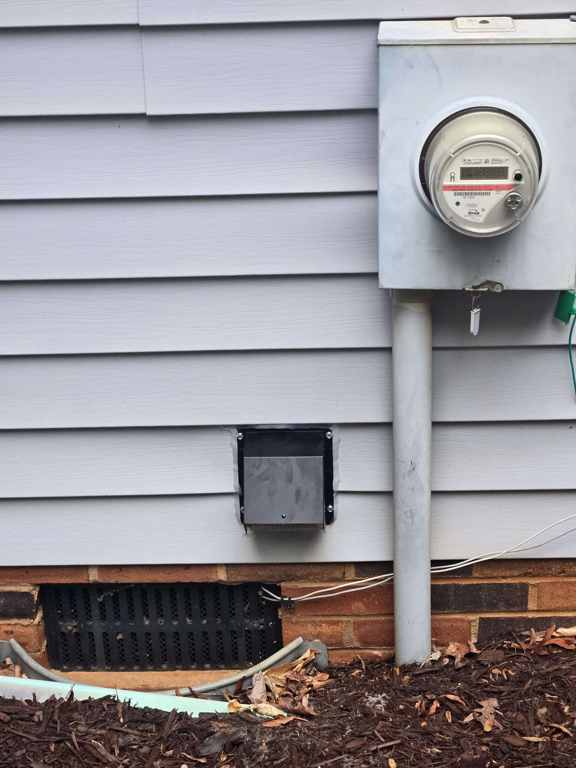 House exterior with electric meter, vent, and mulch bed along gray siding