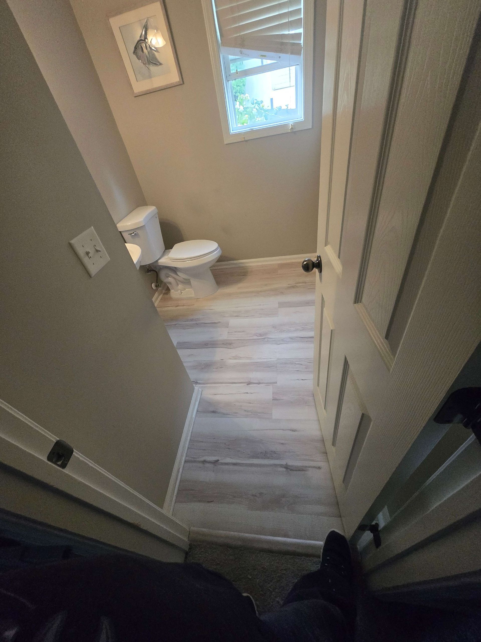 View into a small beige bathroom with toilet, window, and light wood floor from a doorway