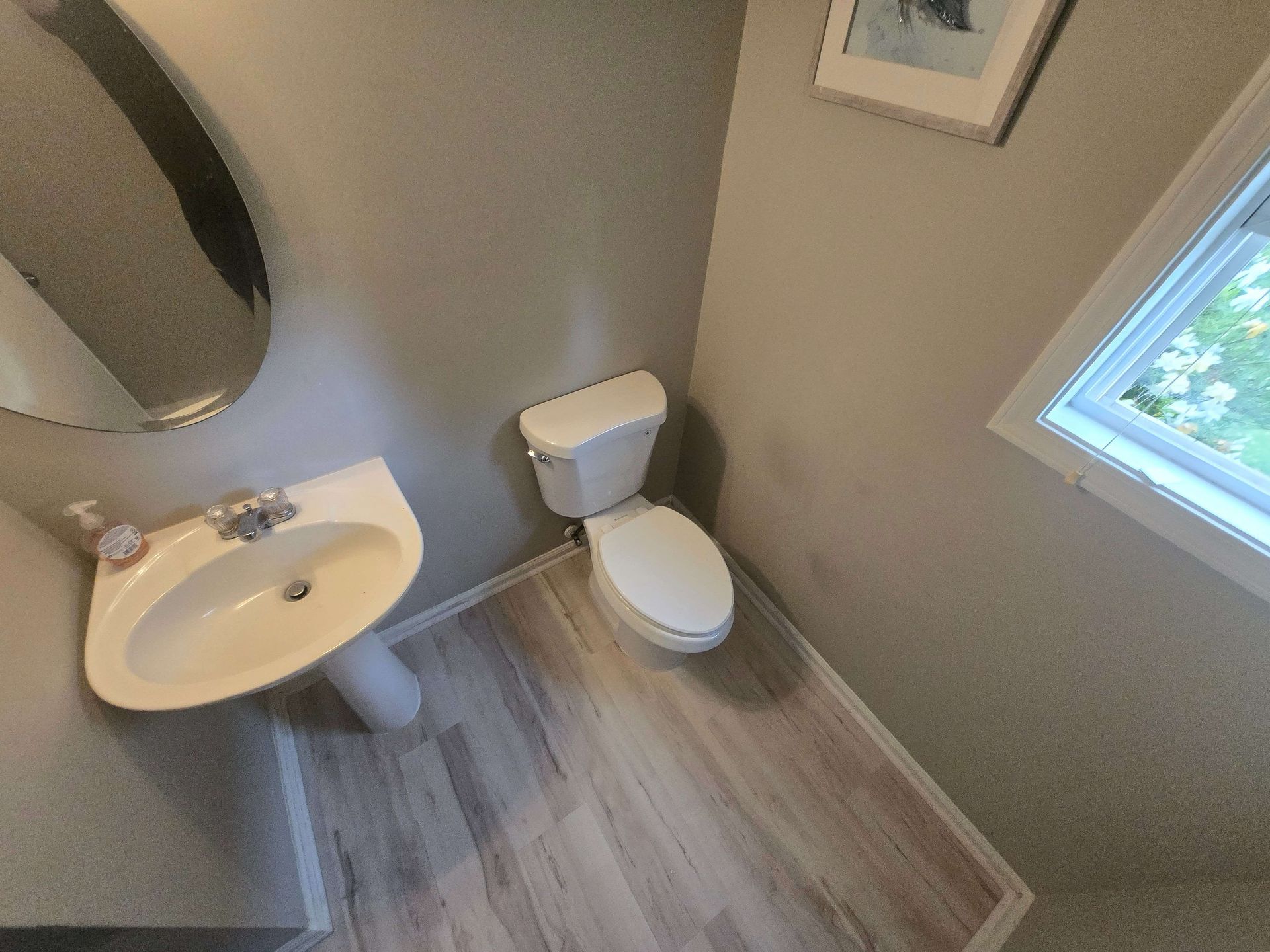 Small bathroom with white sink and toilet, gray walls, wood-look floor, and a window.