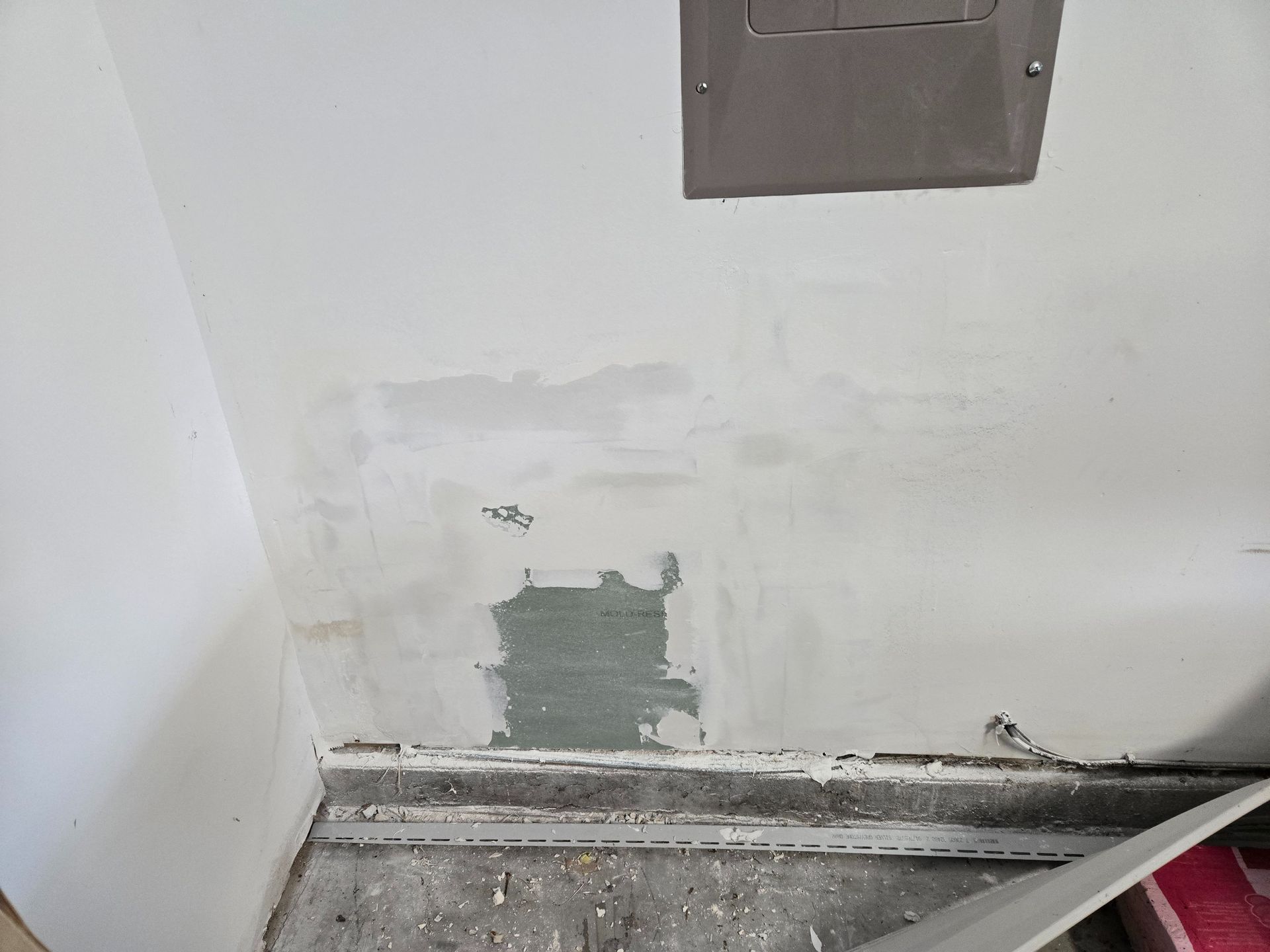 Damaged wall with peeling paint and exposed patch above a baseboard, with a metal panel near the top.