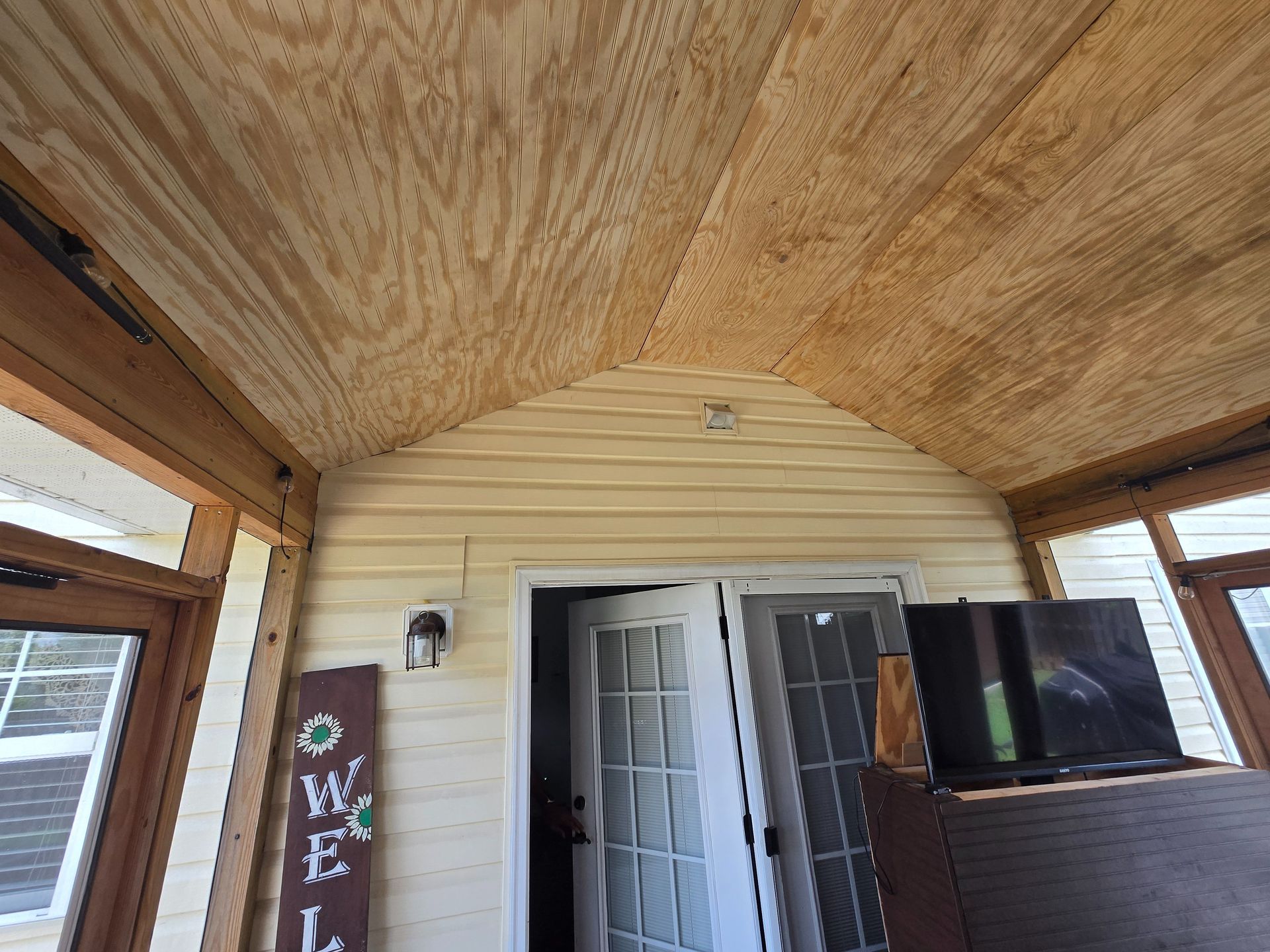 Covered porch with a wood-paneled ceiling, open front door, and railing beside windows and a TV