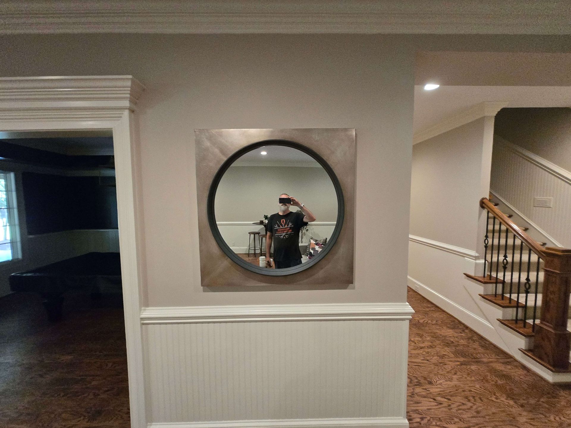 Hallway with round mirror, white wainscoting, and staircase beside wood floor; person visible in mirror reflection
