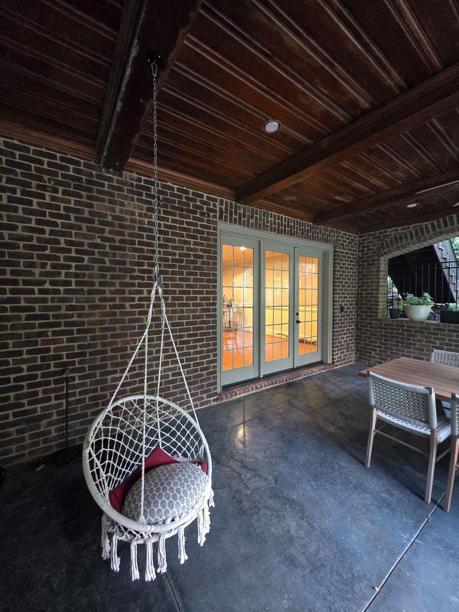 Covered brick patio with wood ceiling, hanging chair, lit French doors, and a small table and chairs.