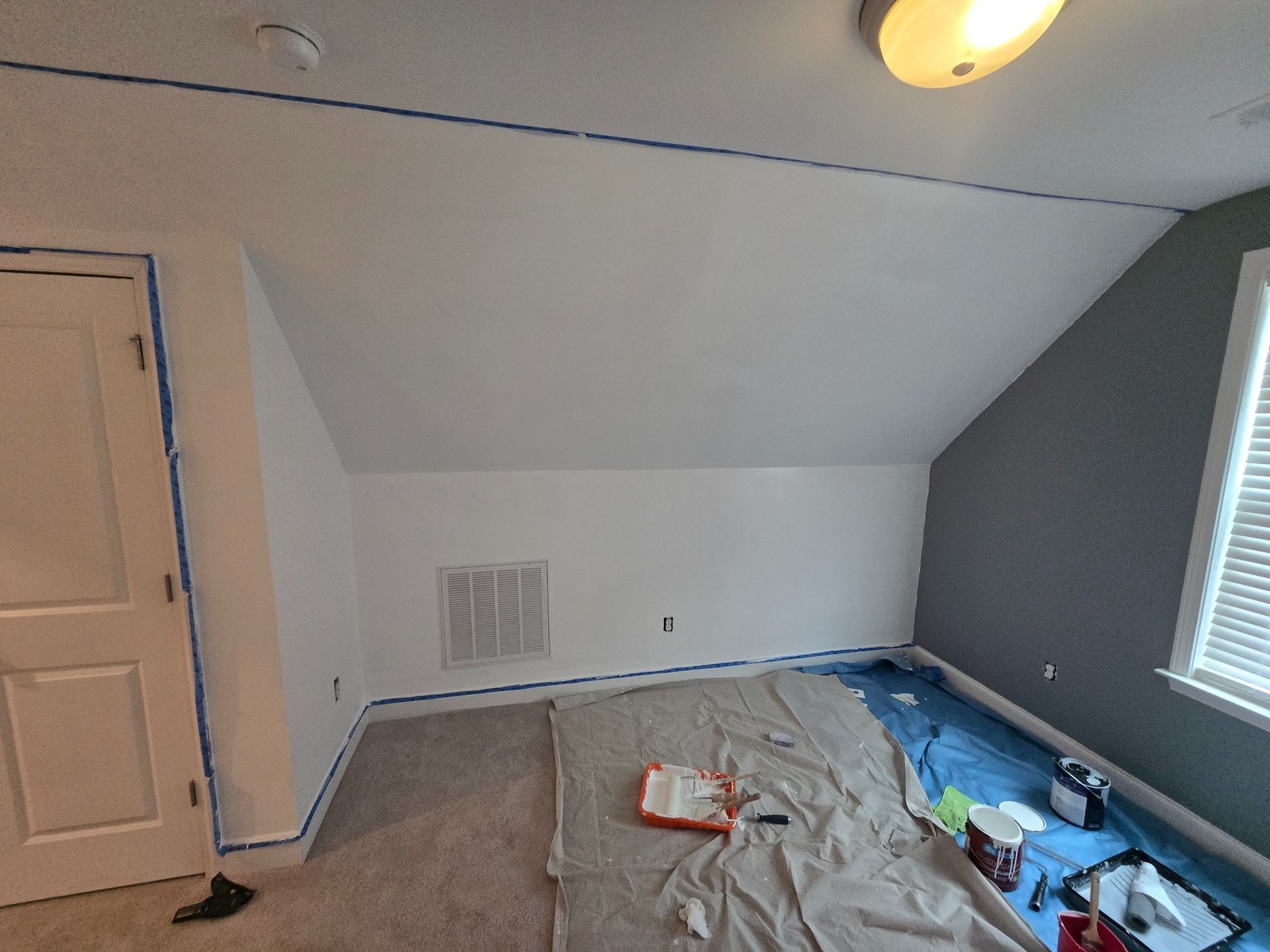 Partially painted attic bedroom with sloped ceiling, bare walls, and floor covered in drop cloths and paint supplies