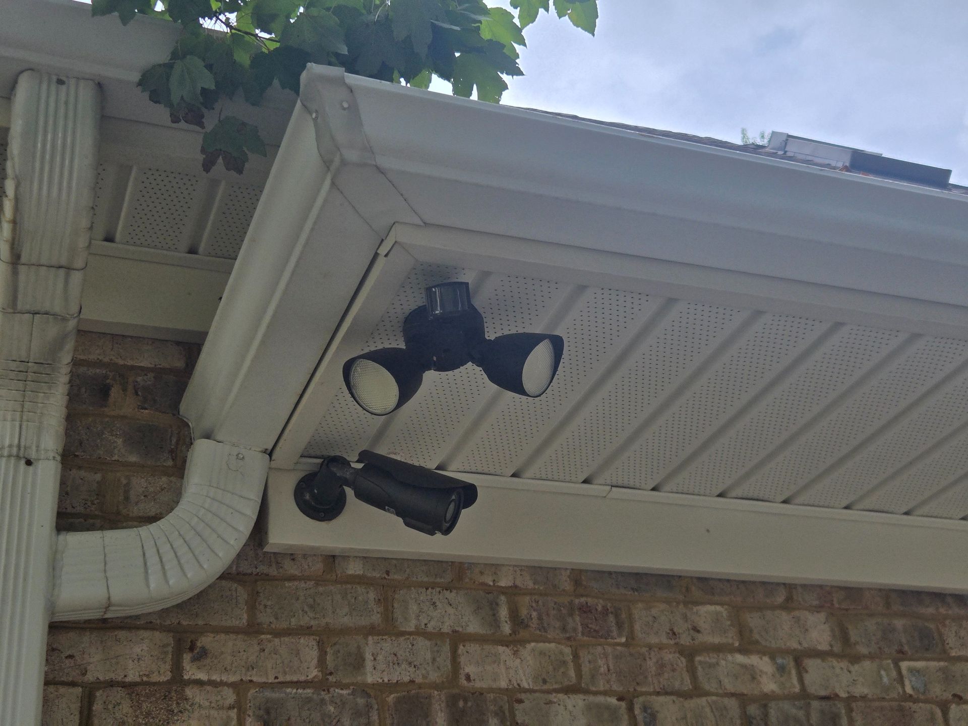 Two black security lights mounted under a white house eave above a brick wall