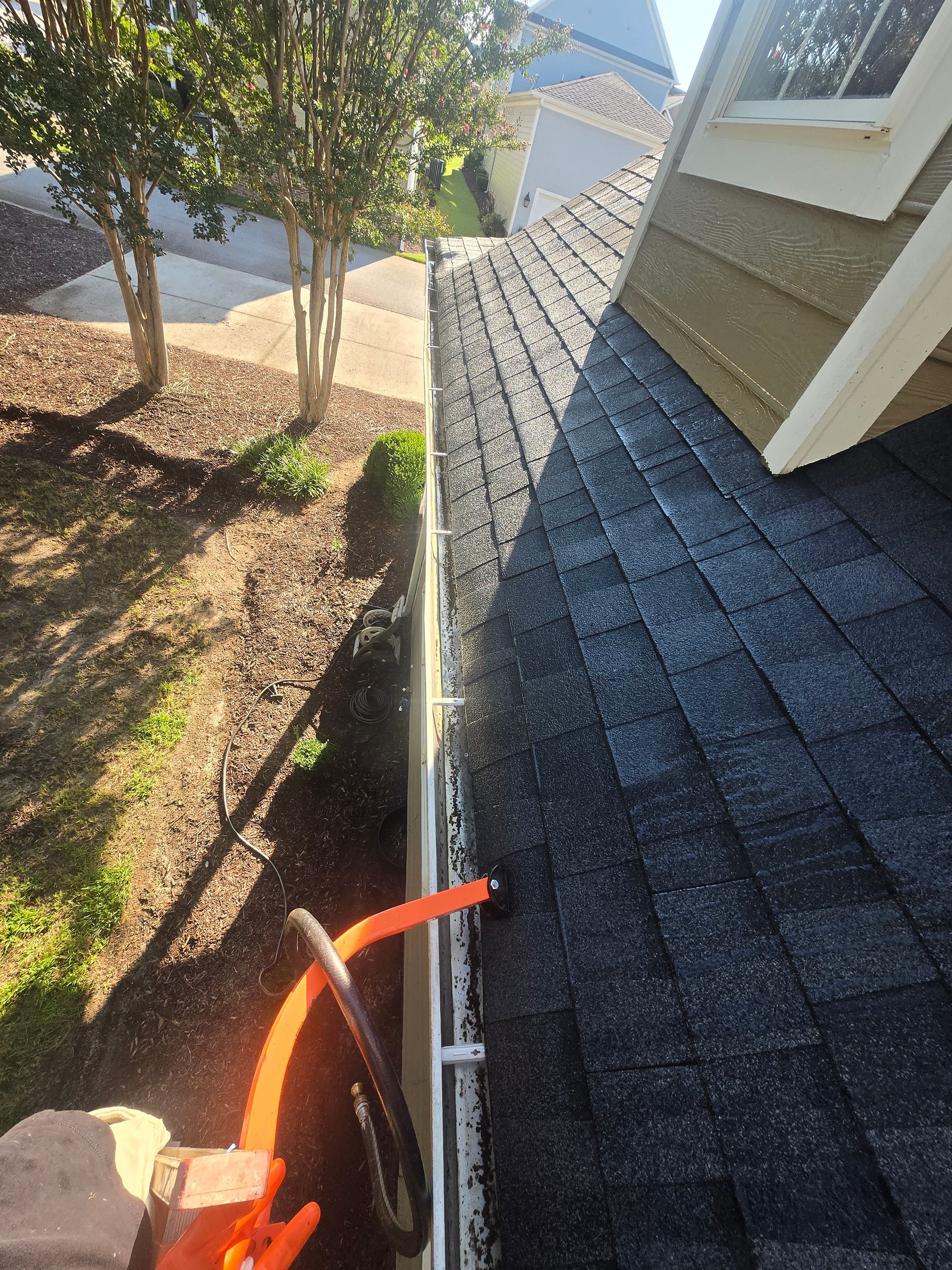Roof edge with dark shingles, gutter, and orange hose beside a house and trees in a yard