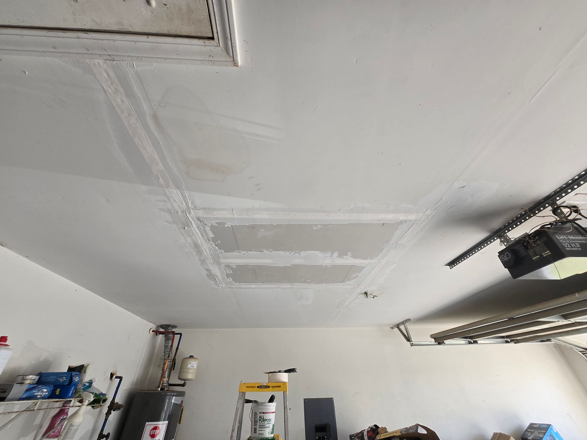 Garage ceiling with white patched drywall and exposed utility fixtures above shelves and a ladder