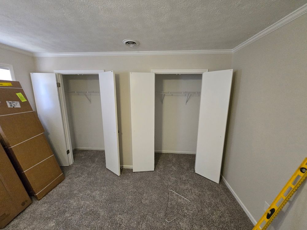 Bedroom with two white closets, carpet, and open closet doors. Cardboard boxes and a level are in the room.
