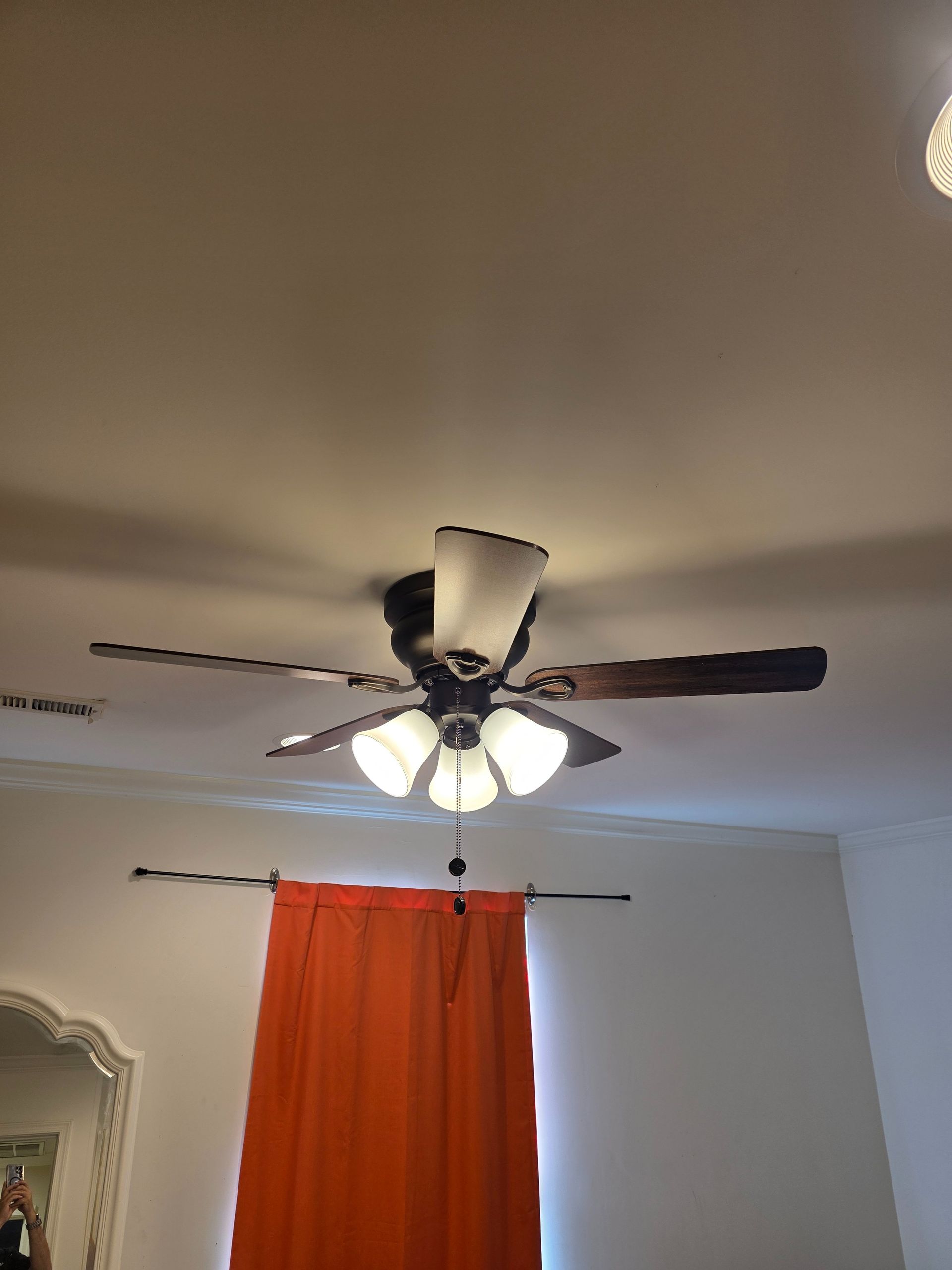 Ceiling fan with lights above an orange curtain in a room with a mirror and white wall.