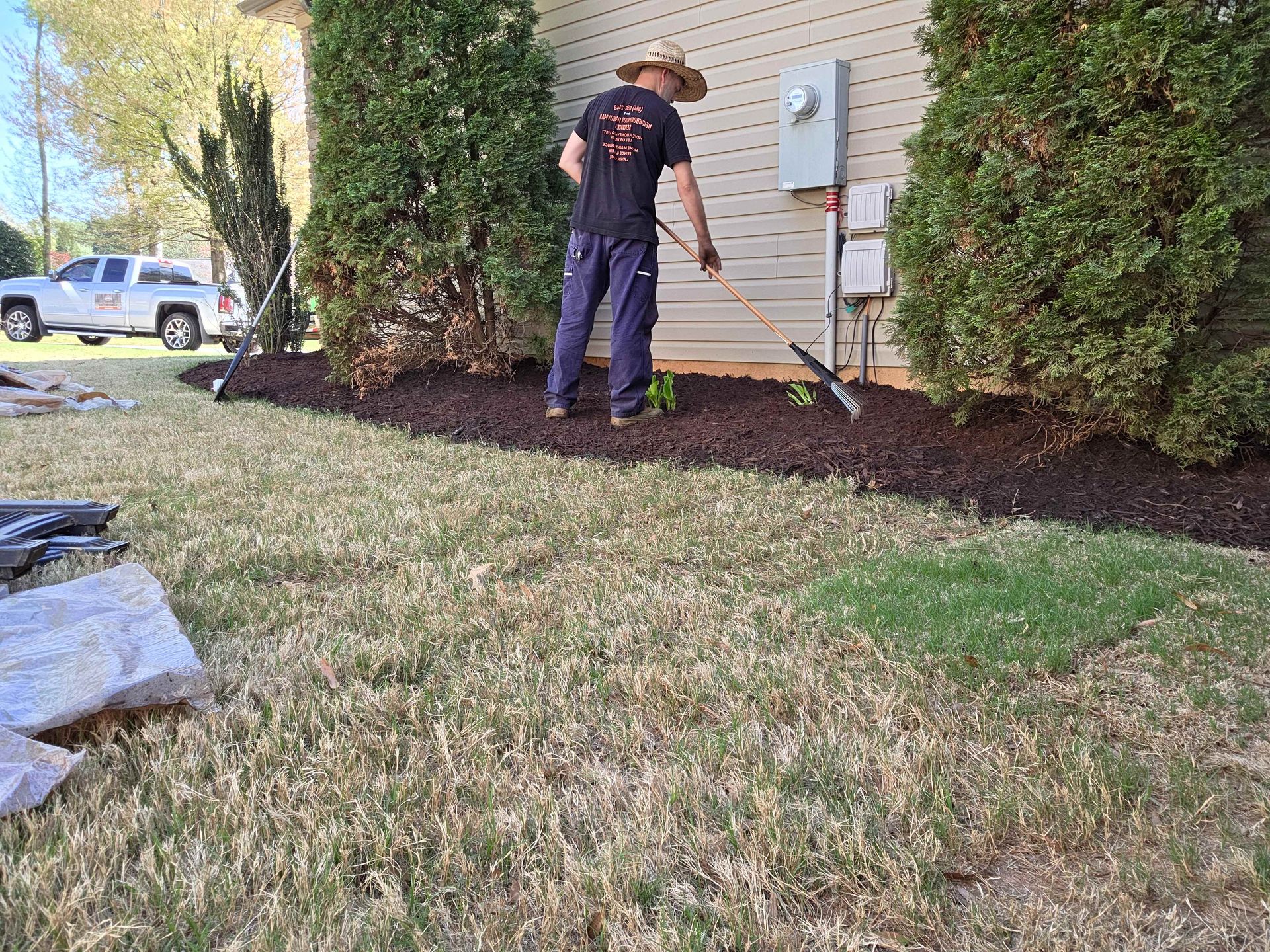 Worker spraying mulch along house foundation beside hedges and electrical panel