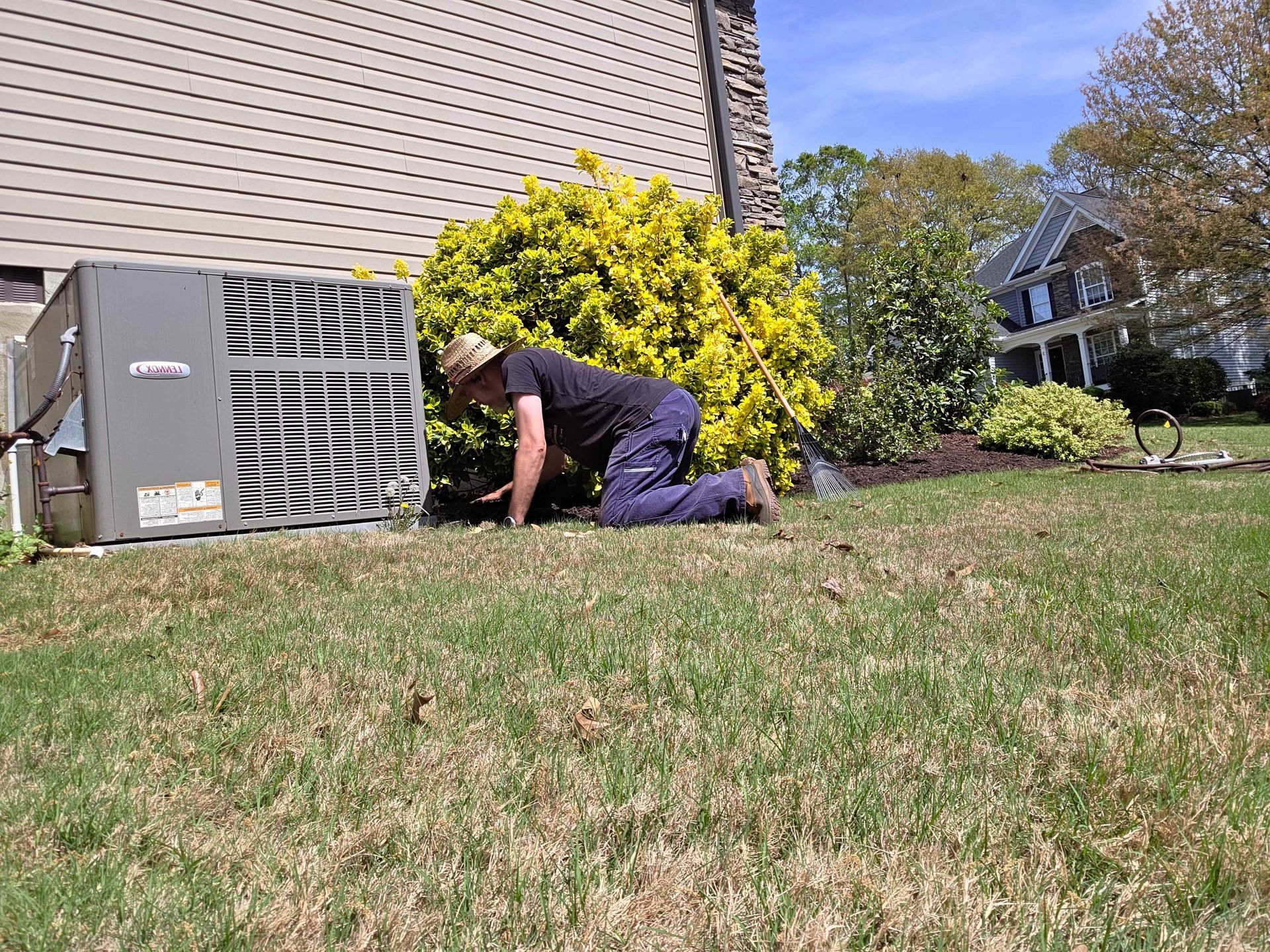 Person kneeling on grass beside a house, working near an outdoor air conditioner unit.