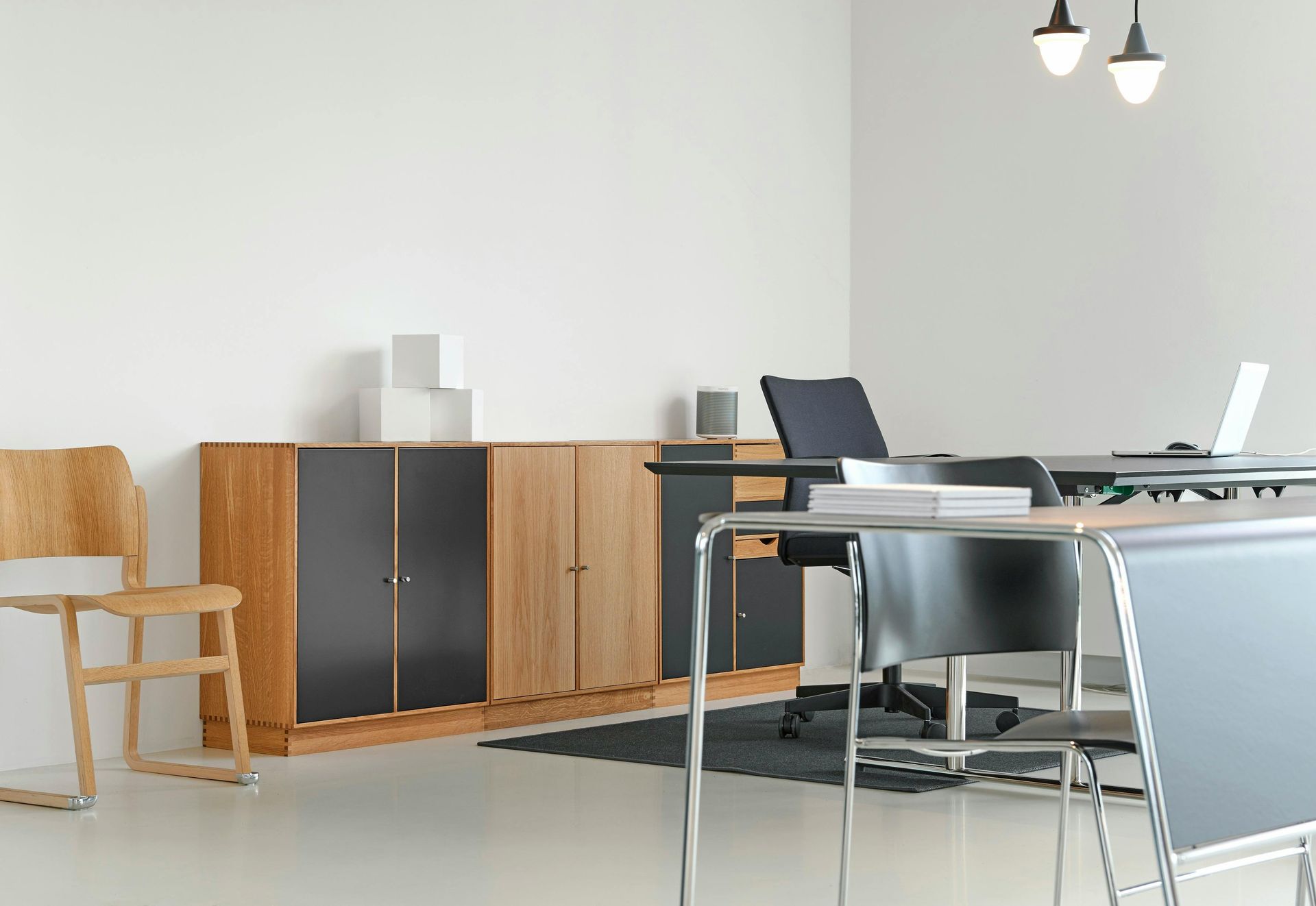 An office with wooden cabinets, desk, chairs, and a laptop against a white wall, under pendant lights.