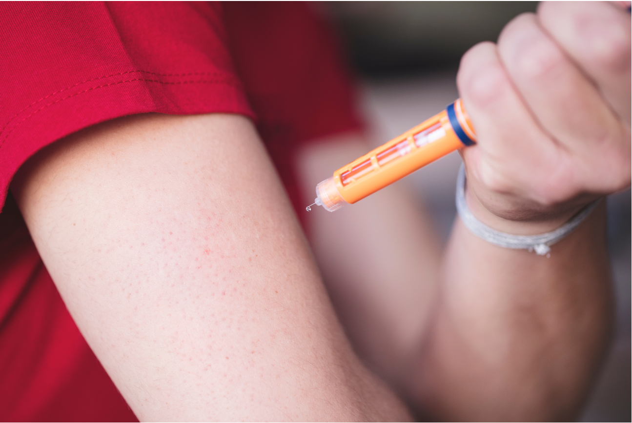 Hand holding an orange insulin pen near an upper arm in a red shirt
