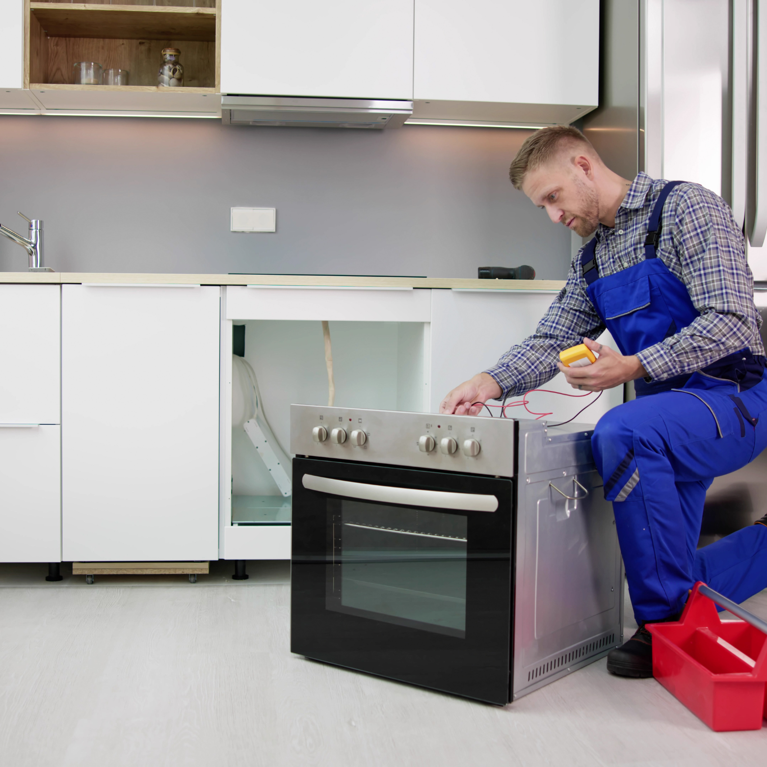 A man in blue overalls is working on an oven in a kitchen.