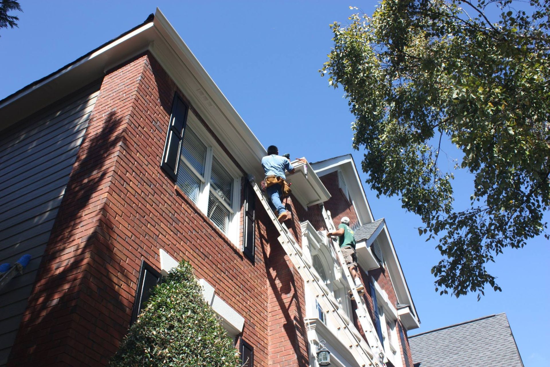 Two workers on extension ladders install gutters on the brick exterior of a multi-story home under a clear blue sky.