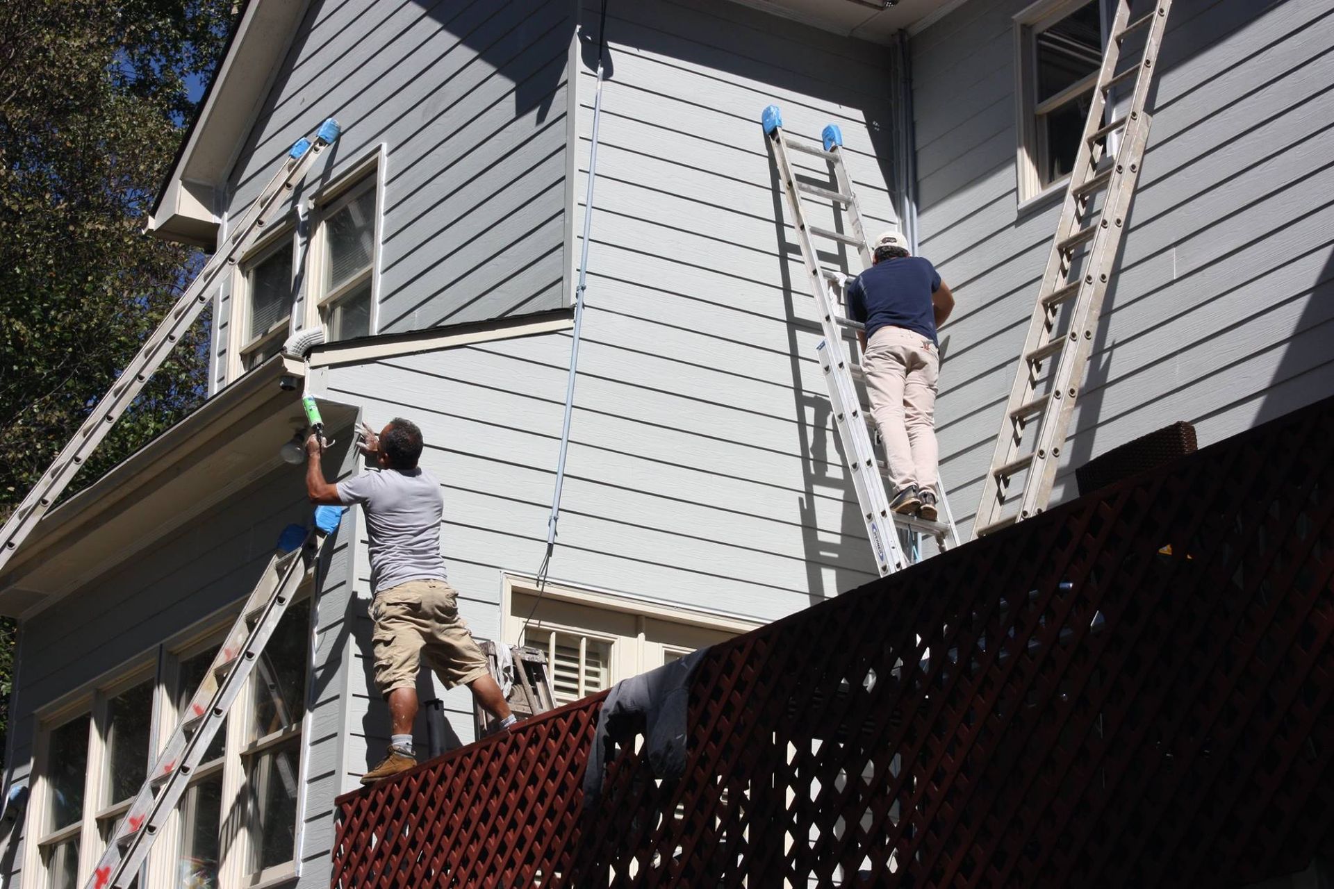 Two people use ladders to work on the exterior gray siding of a two-story home.