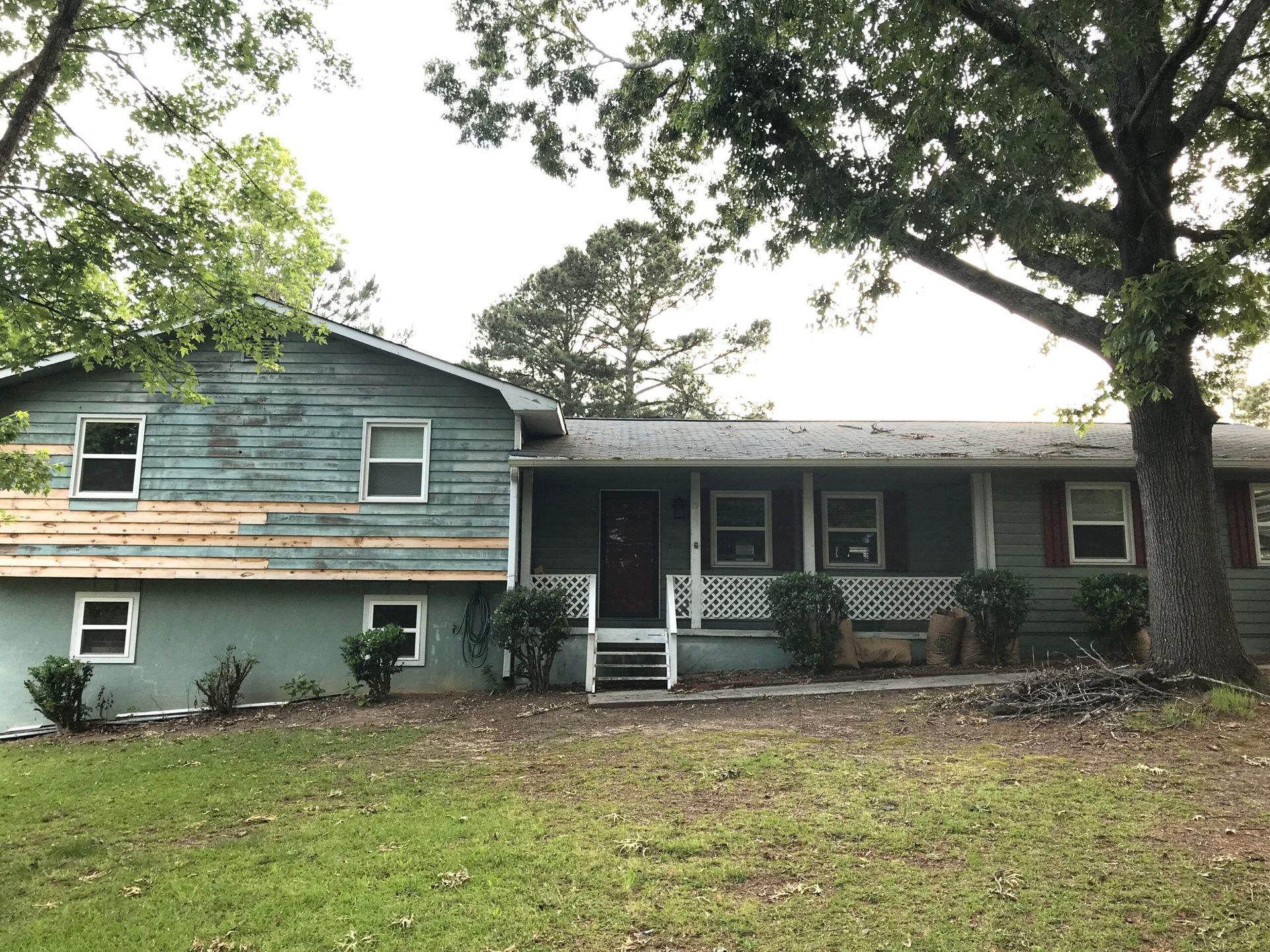 A split-level home with faded blue-green siding and an exposed wood section, featuring a front porch and large oak trees.