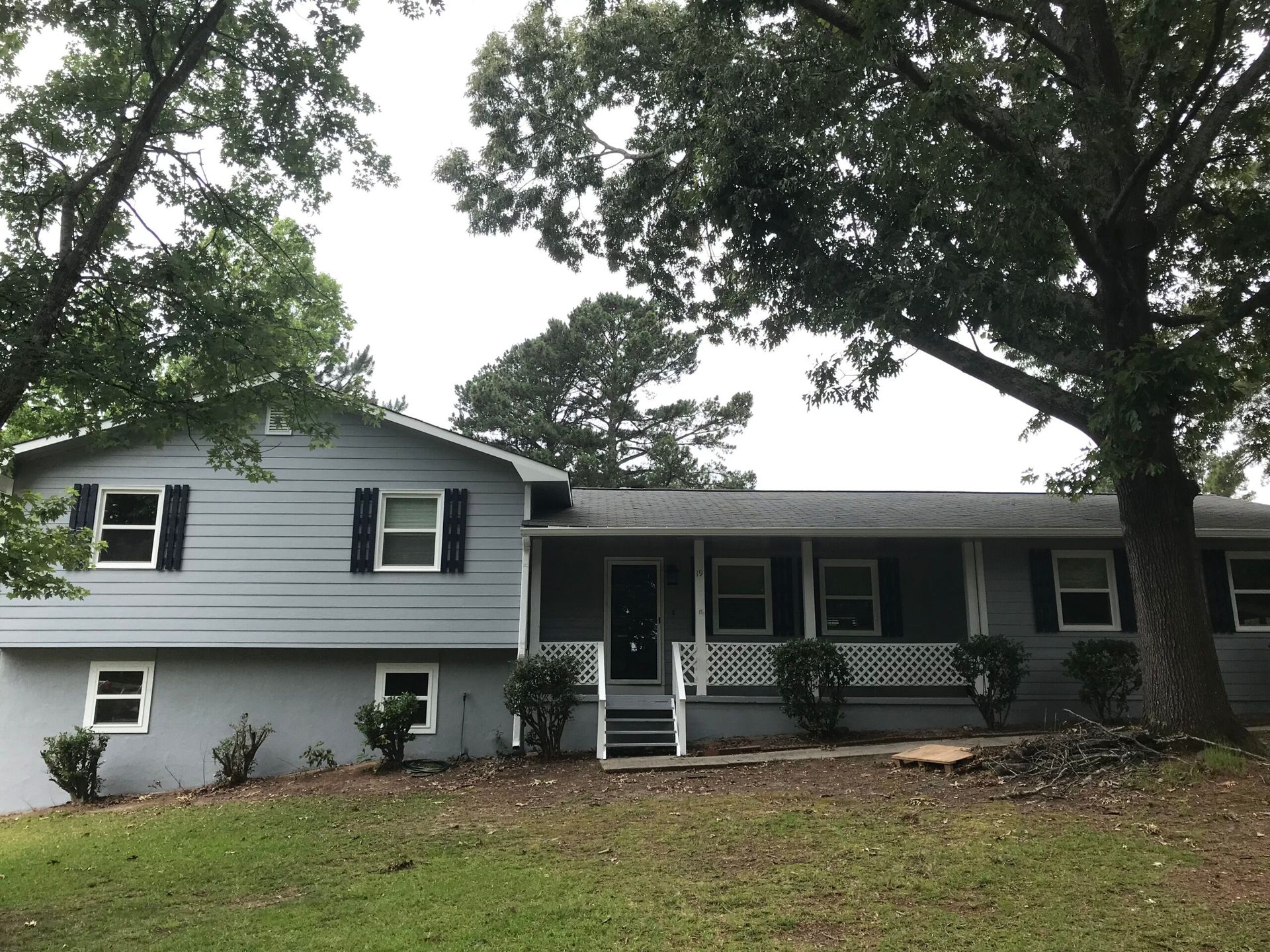 A two-story, gray, horizontal-sided home with a covered porch and front stairs, surrounded by trees and a grassy yard.